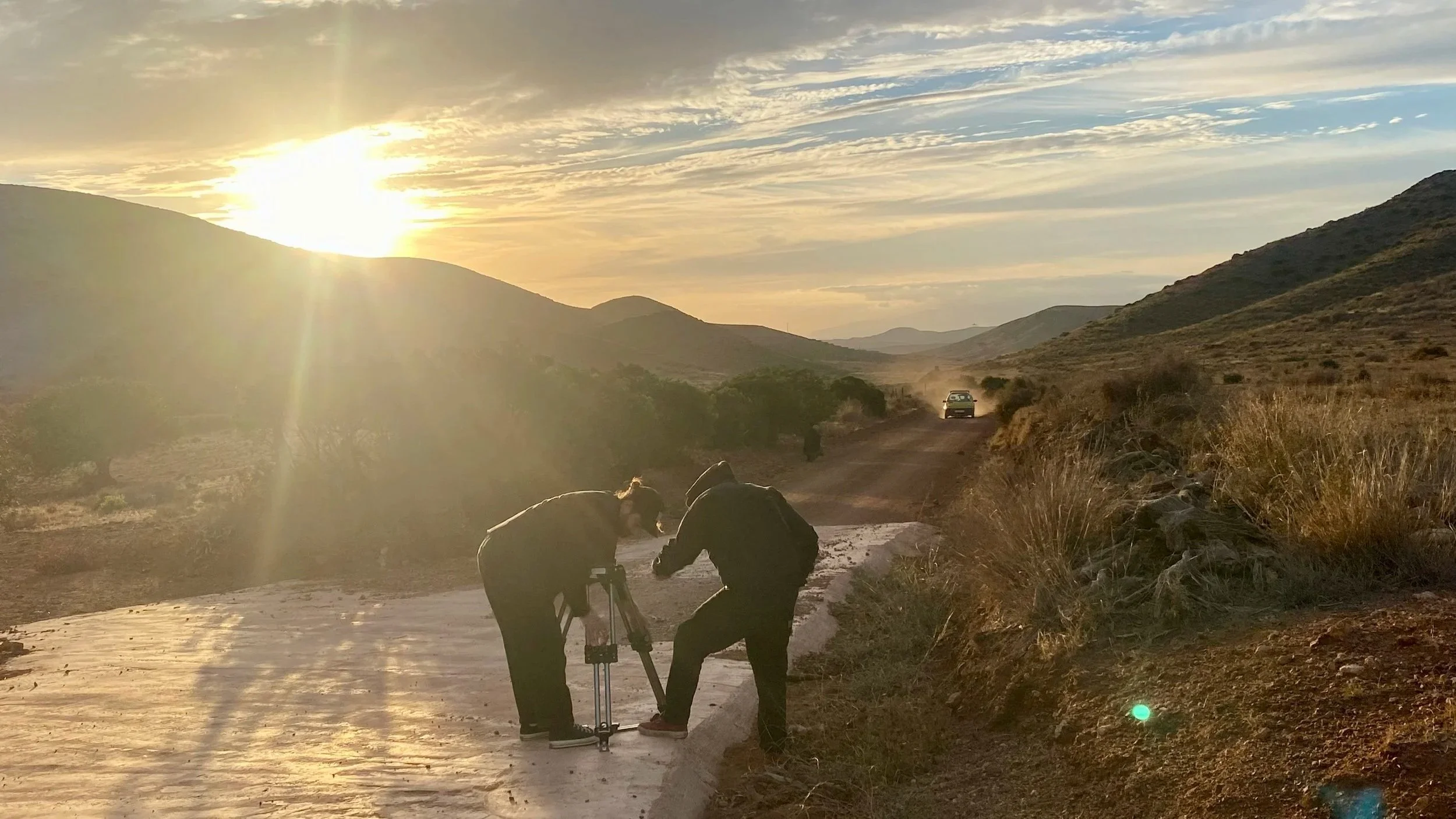 Two people working on camera equipment in a desert landscape during sunset, with a car driving on a dirt road in the background.