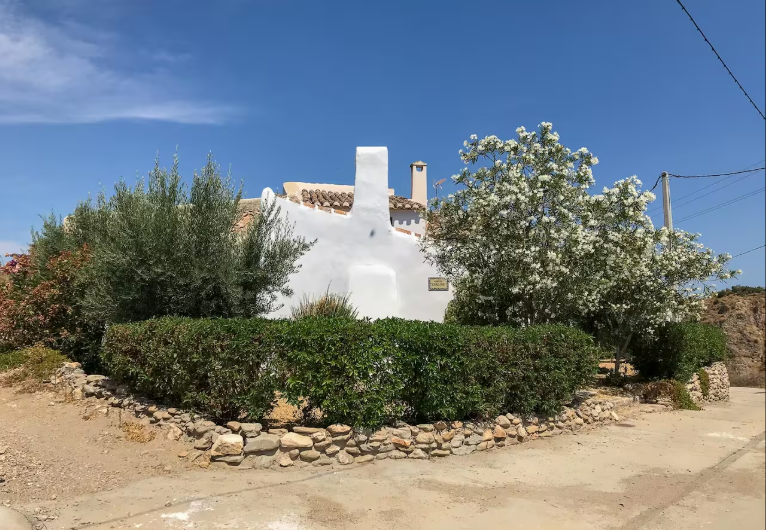 White house with a tiled roof, surrounded by green bushes and flowering trees, under a clear blue sky.