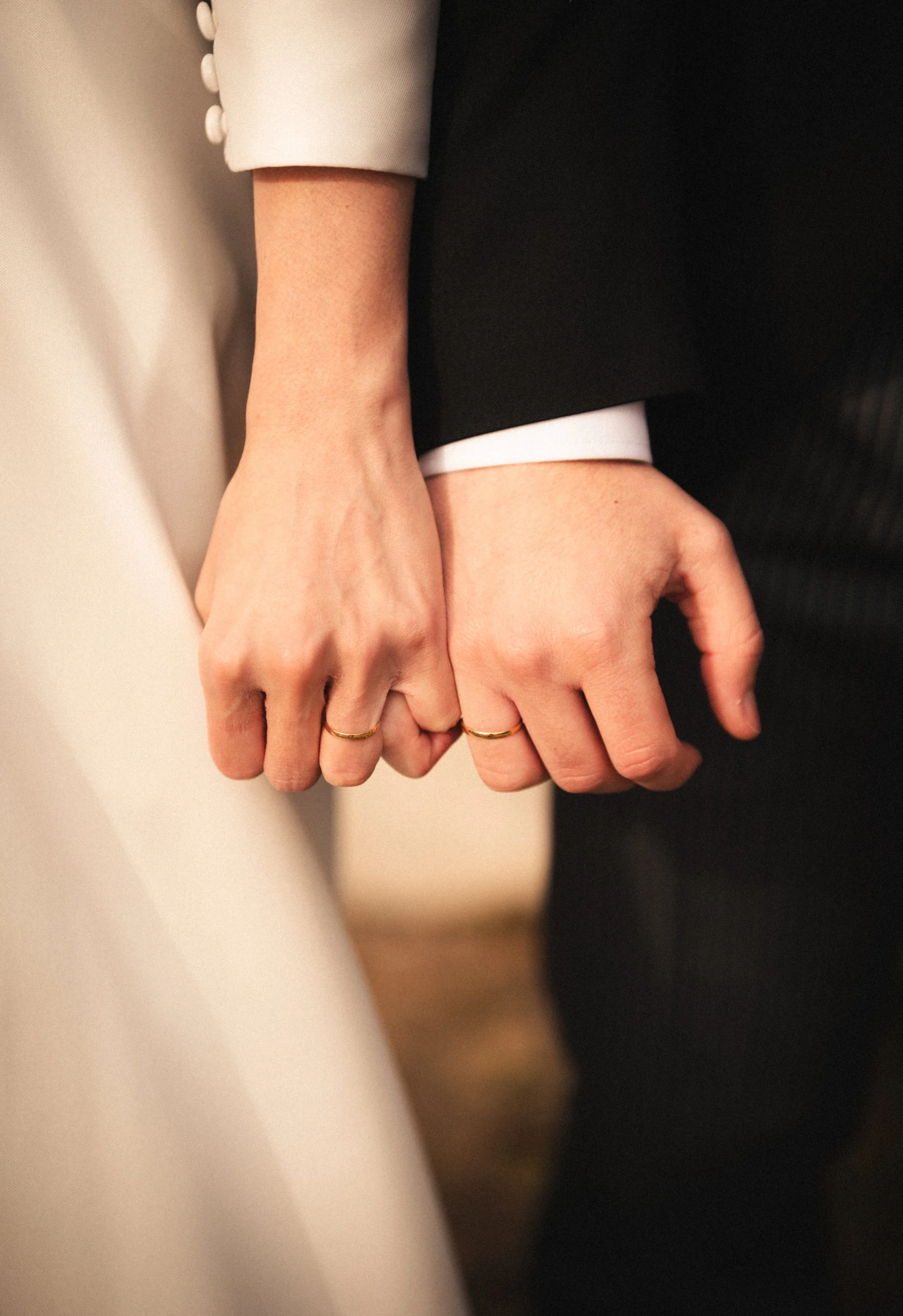 Close-up of two hands holding each other, both wearing rings and dressed in formal attire. One hand wears a white sleeve with buttons, and the other a black sleeve with a white cuff.