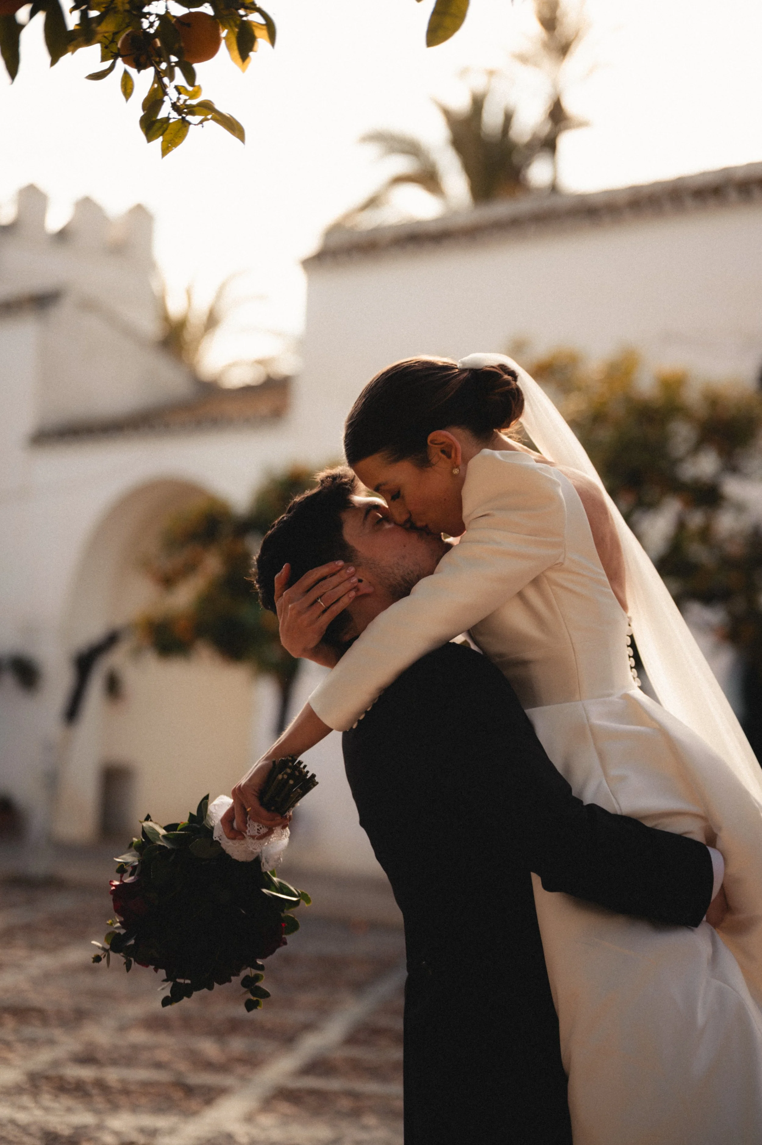 A couple shares a romantic kiss outdoors during sunset, with the woman dressed in a white dress and veil, and the man in dark attire. The woman is perched on the man's shoulders and holding a bouquet of flowers.