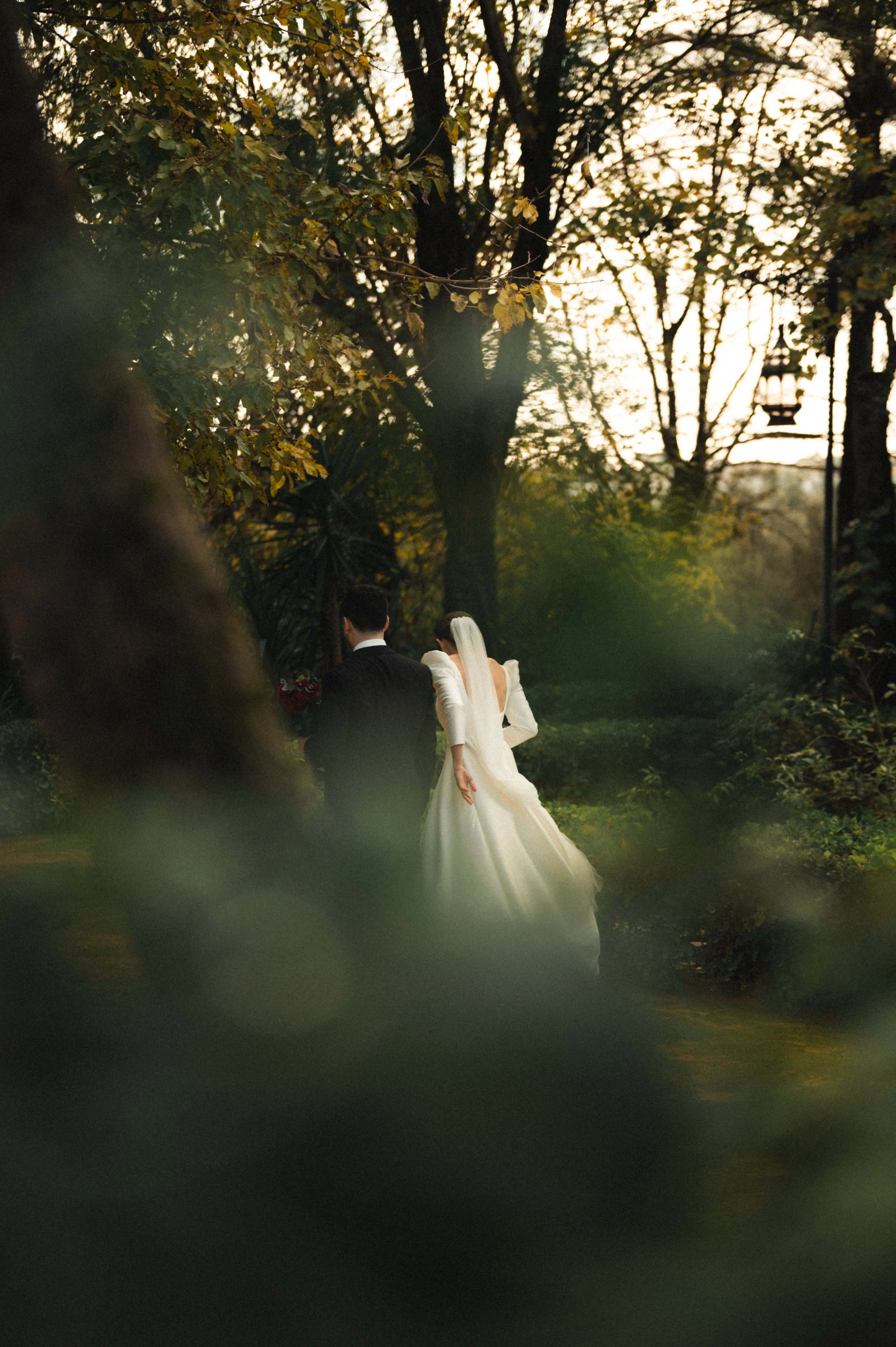 A bride and groom walking through a wooded outdoor area during sunset, with the bride wearing a white wedding dress and the groom in a dark suit.