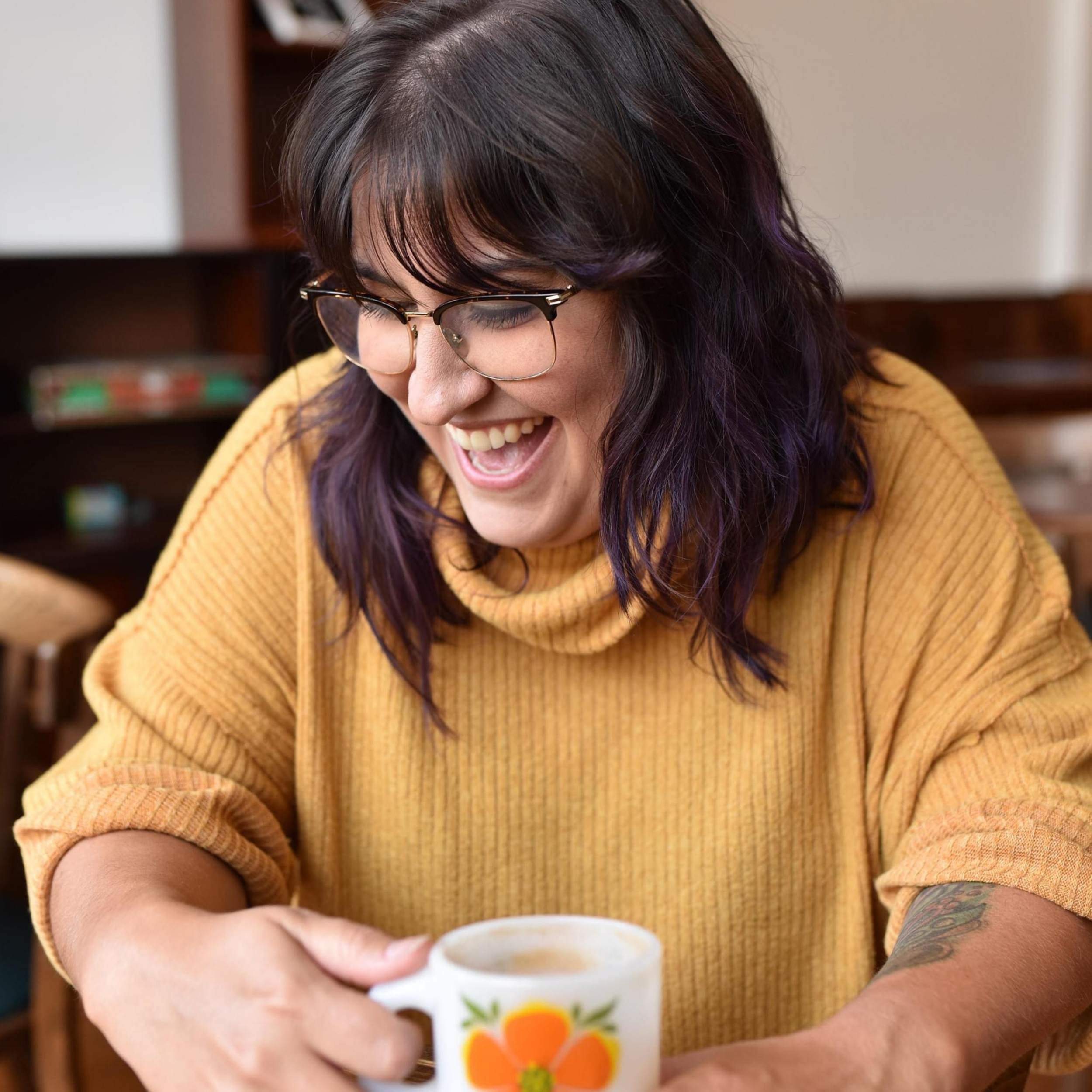 A woman with dark brown hair with purple highlights and glasses holding a mug with orange flowers design.