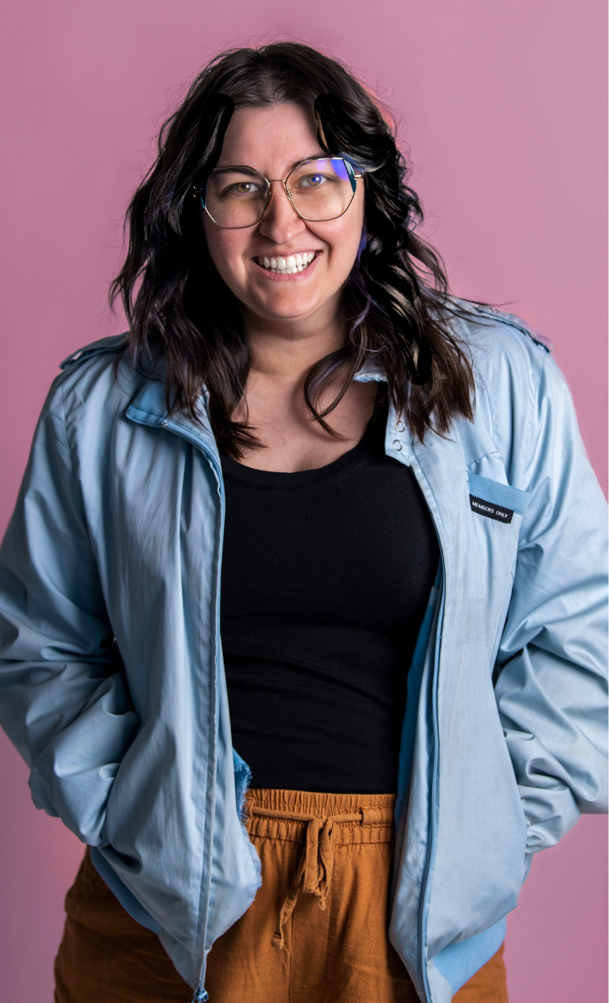 A woman with dark shoulder-length curly hair wearing glasses, a light blue jacket over a black top, and brown pants with a drawstring, smiling against a pink background.