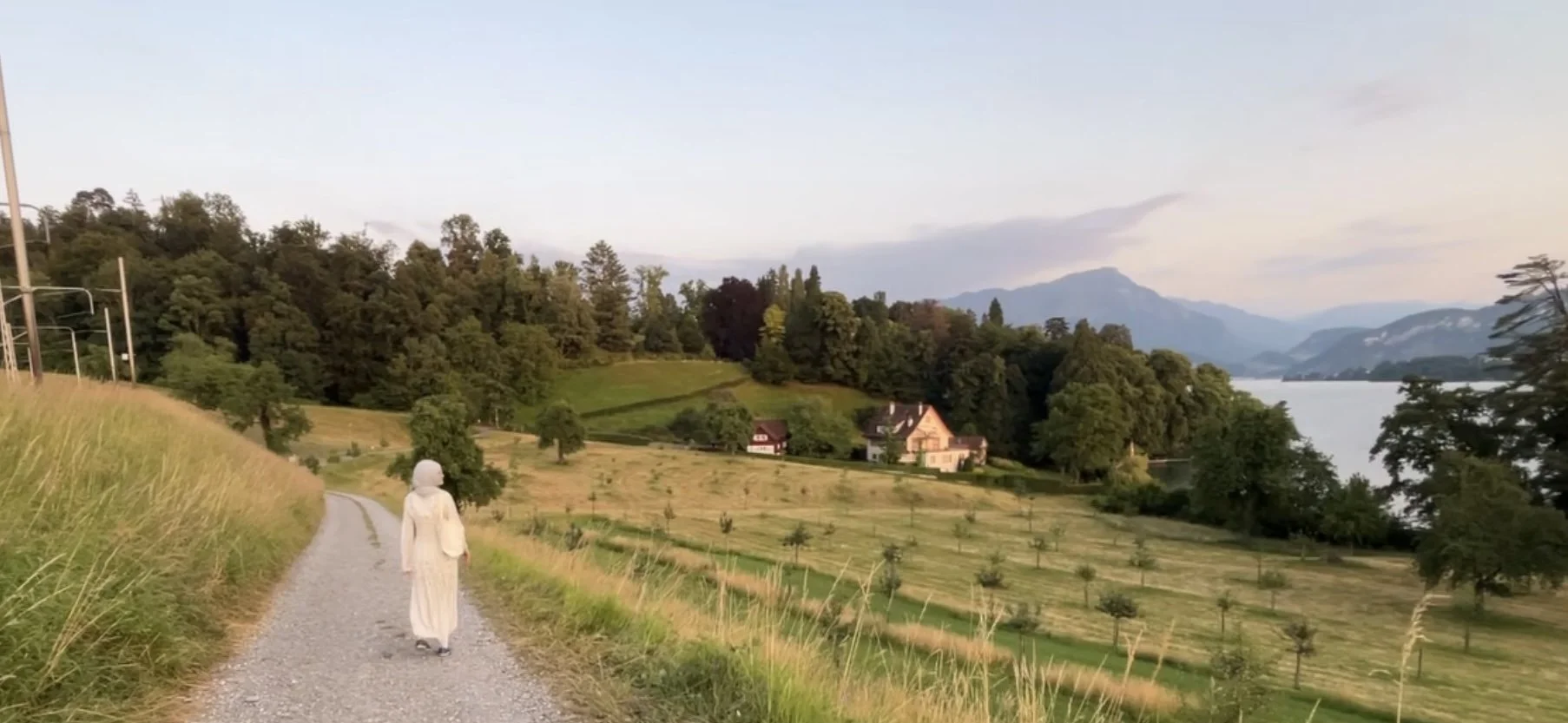 A woman in a white dress walking on a gravel path through a hilly, green landscape; houses among trees, a lake, mountains in the background, and a cloudy sky.