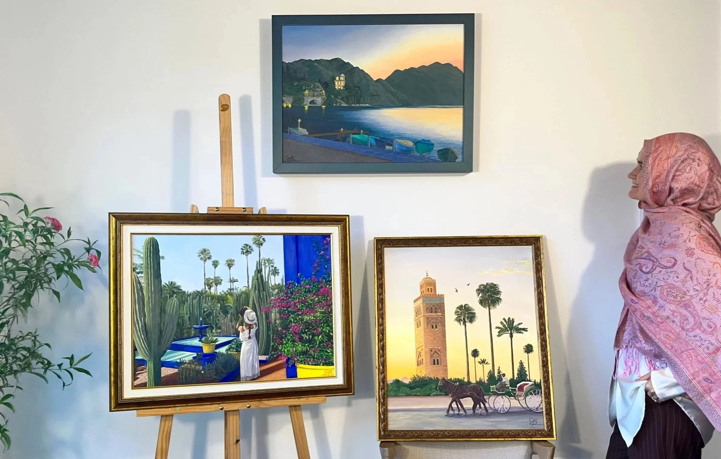 A female artist looking at her three original oil paintings from which she sells her fine art prints. One depicts a serene lakeside sunset with boats, the second is a horse carriage passing in front of a mosque. Lastly a woman in a garden.