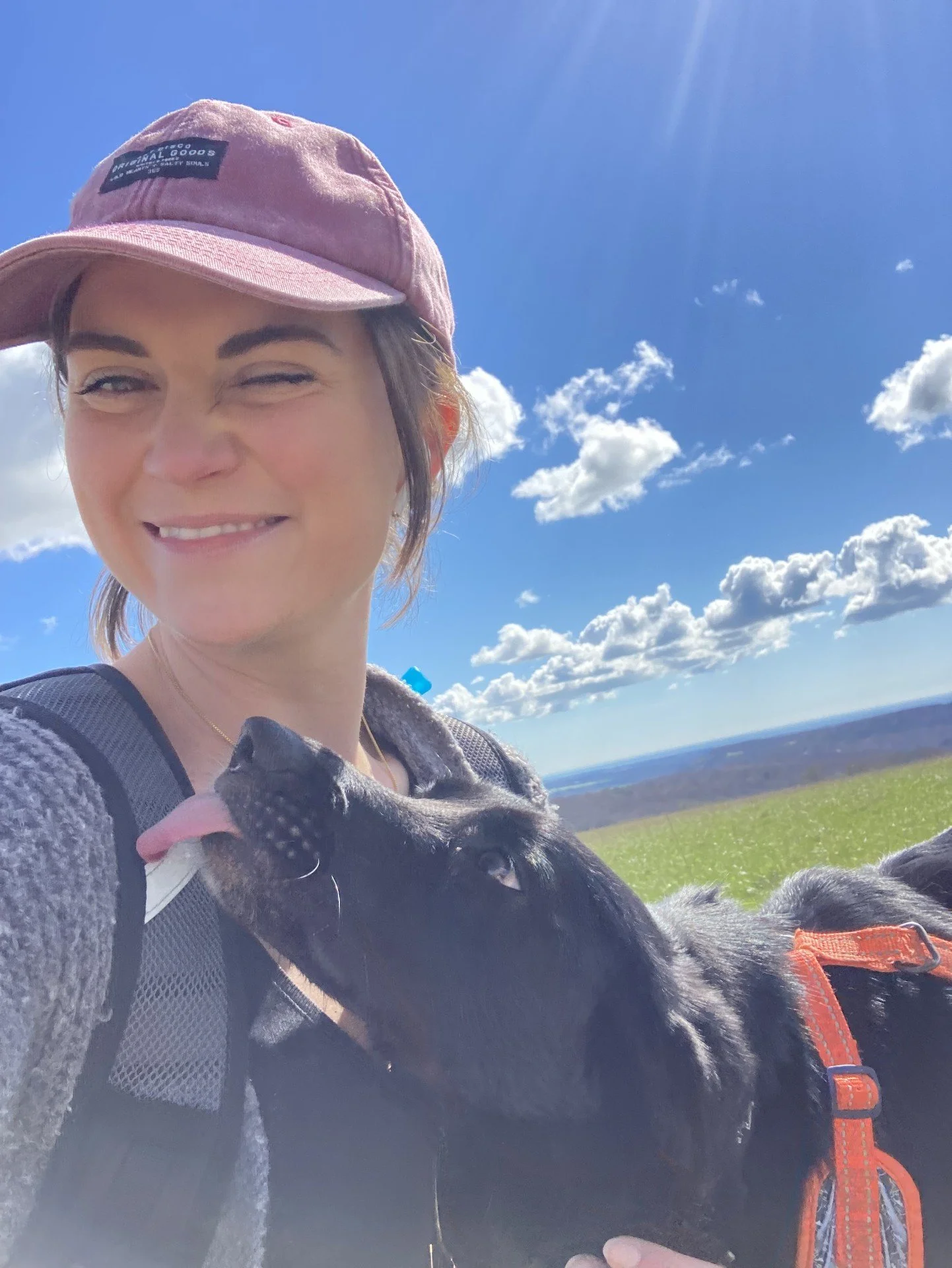 Sophie & Winnie her black dog outdoors on a sunny day. The dog is licking her cheek, and the background features a bright blue sky with scattered clouds and a green landscape.