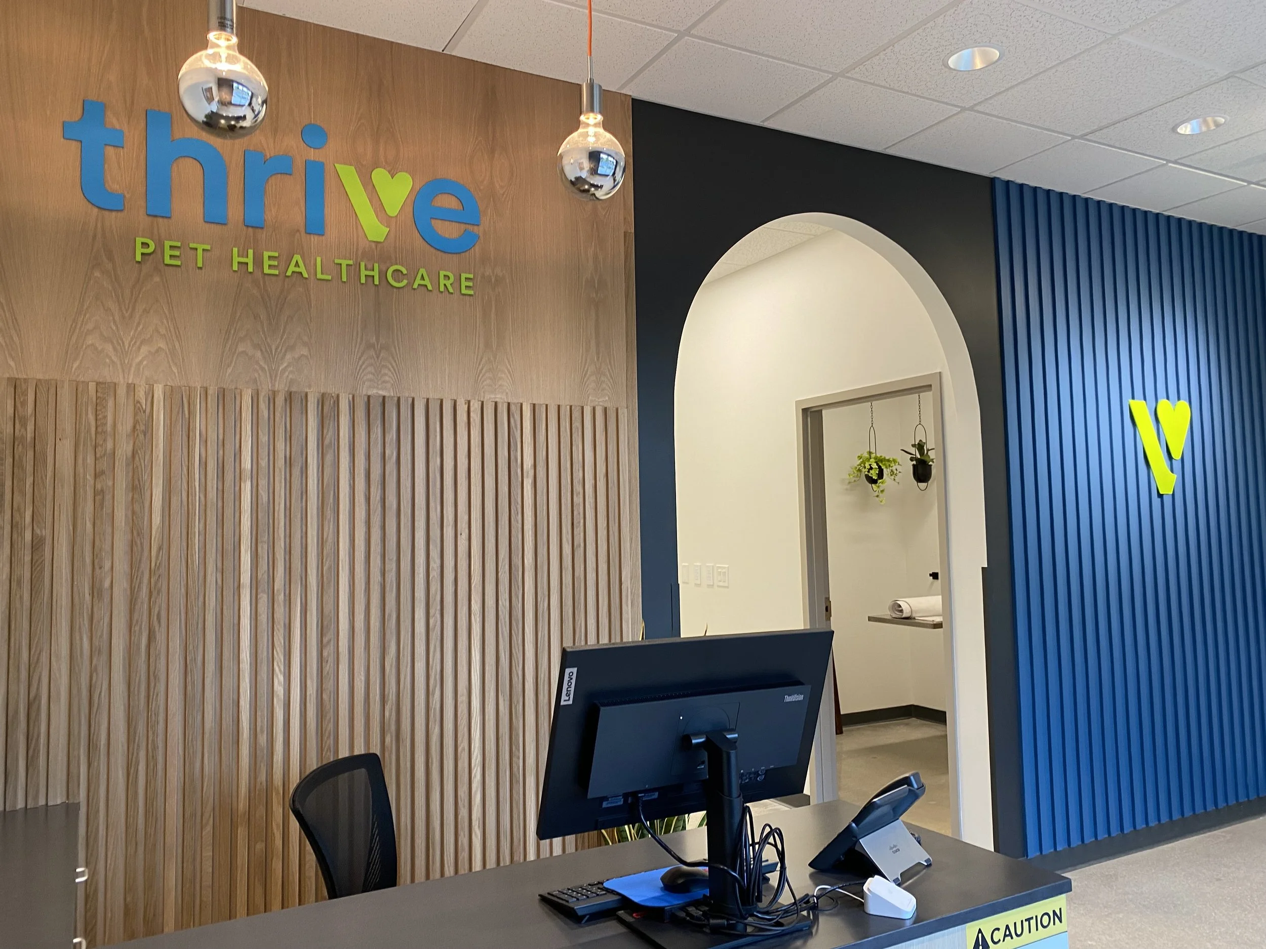 Reception desk at Thrive Pet Healthcare with a computer, phone, and office supplies in front of a wooden wall with Thrive Pet Healthcare sign and two hanging silver lights.