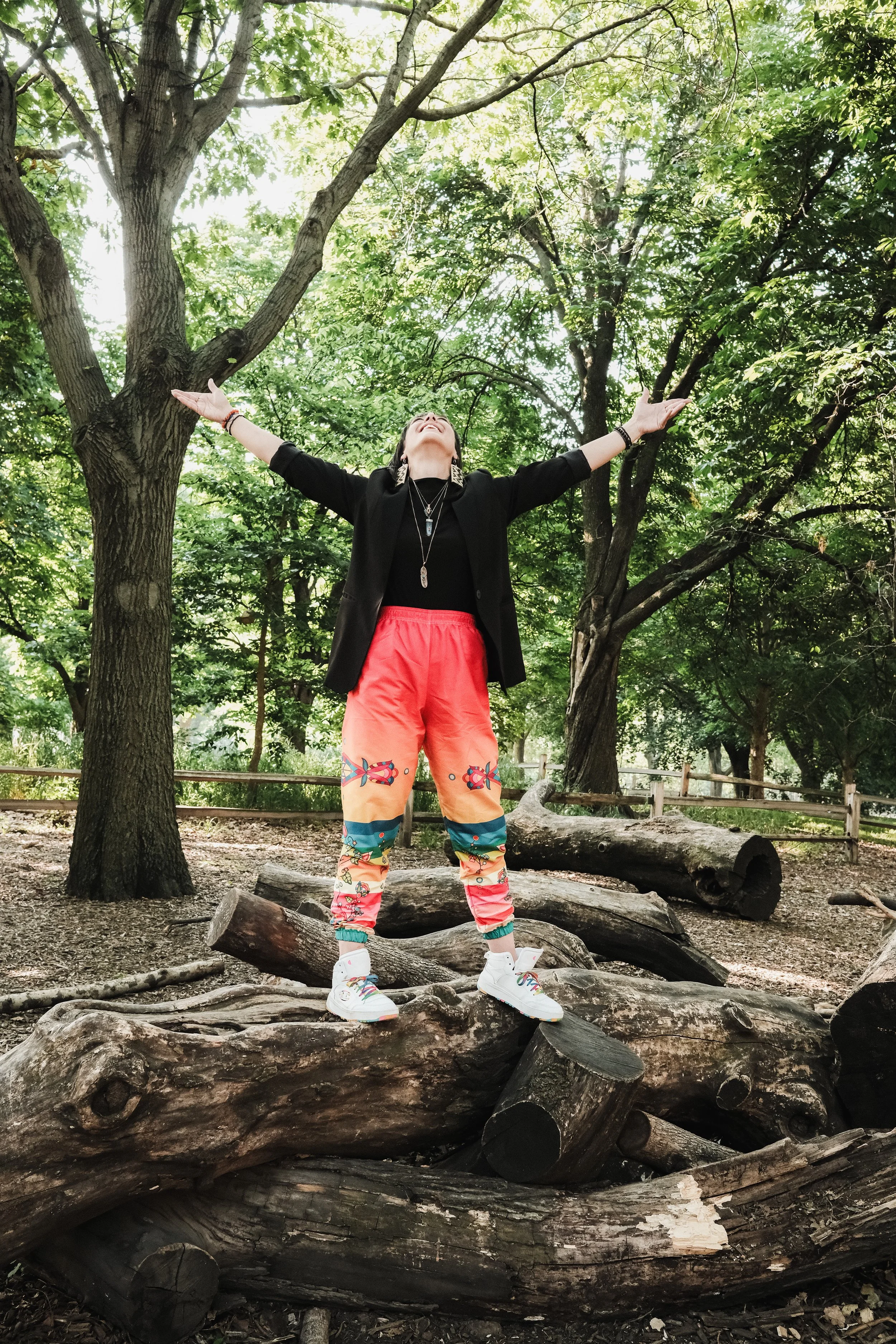 A woman standing on fallen tree logs in a wooded park with arms outstretched, wearing colorful sweatpants, a black jacket, and white sneakers.
