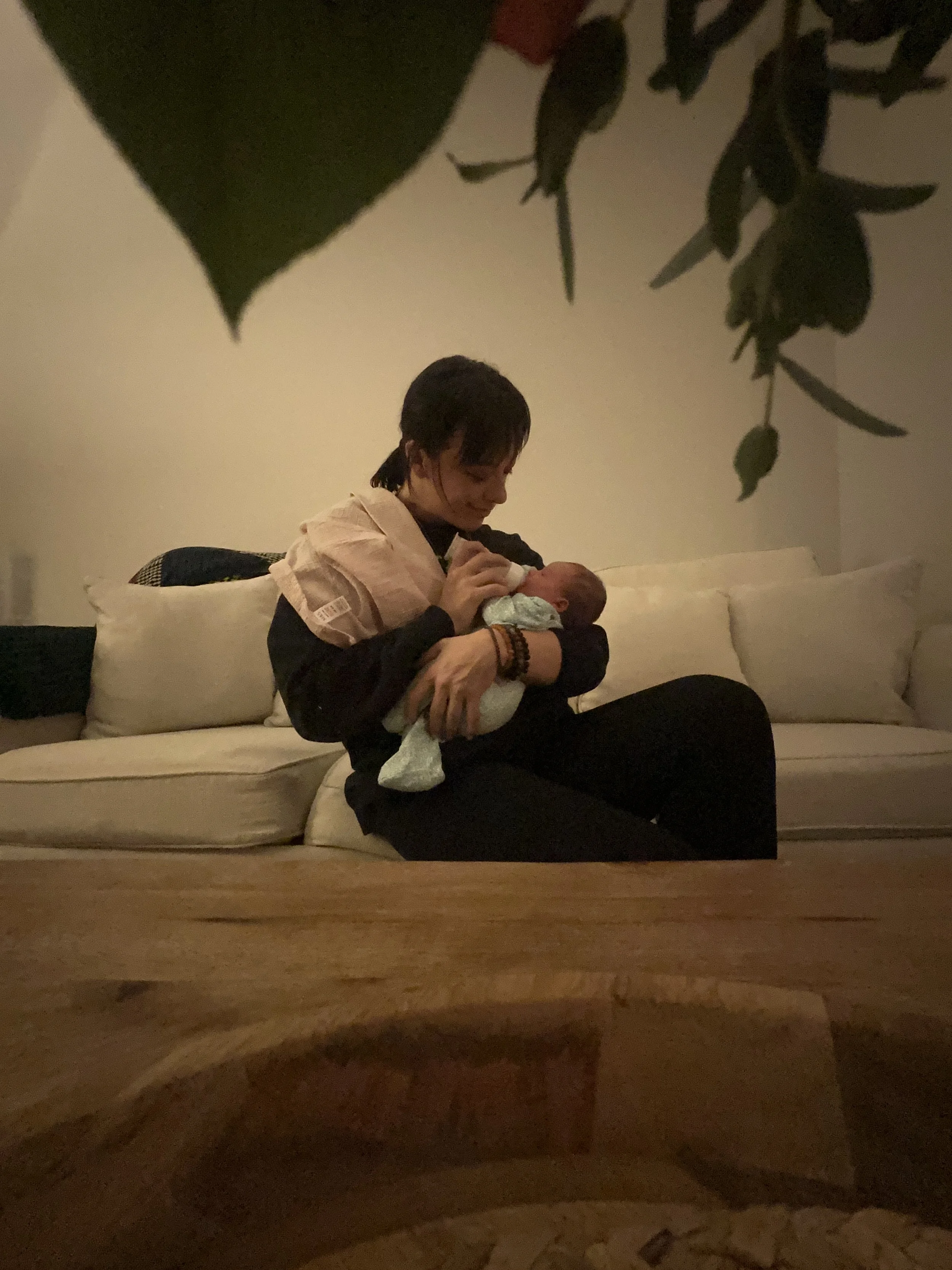 A woman sitting on a couch holding a newborn baby, gazing lovingly at the baby, with a cozy interior setting.