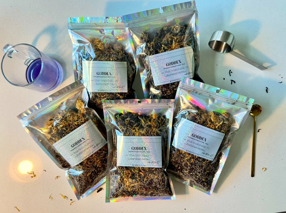 Five resealable bags of dried herbs or tea labeled 'GODDEX Transformation Tea' on a white surface, with a small metal scoop and a purple-tinted glass of water nearby, and loose tea leaves scattered around.