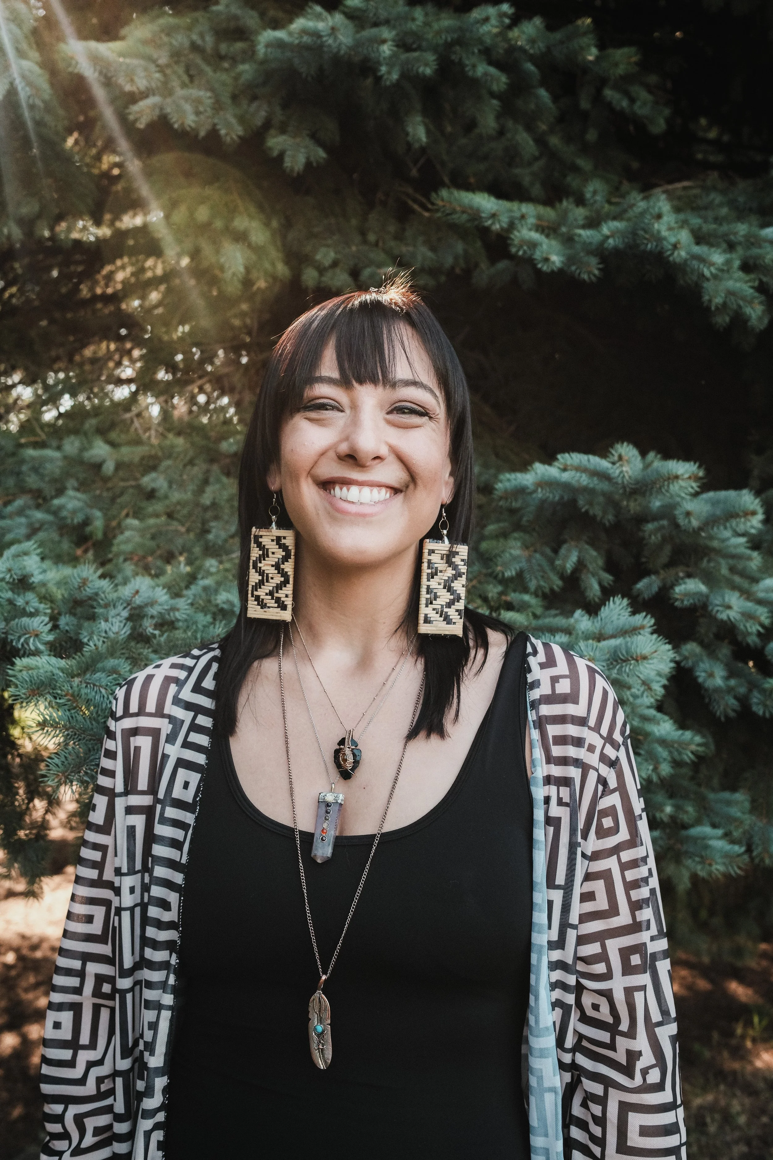 A woman smiling outdoors in front of evergreen trees, sun rays visible in the background.