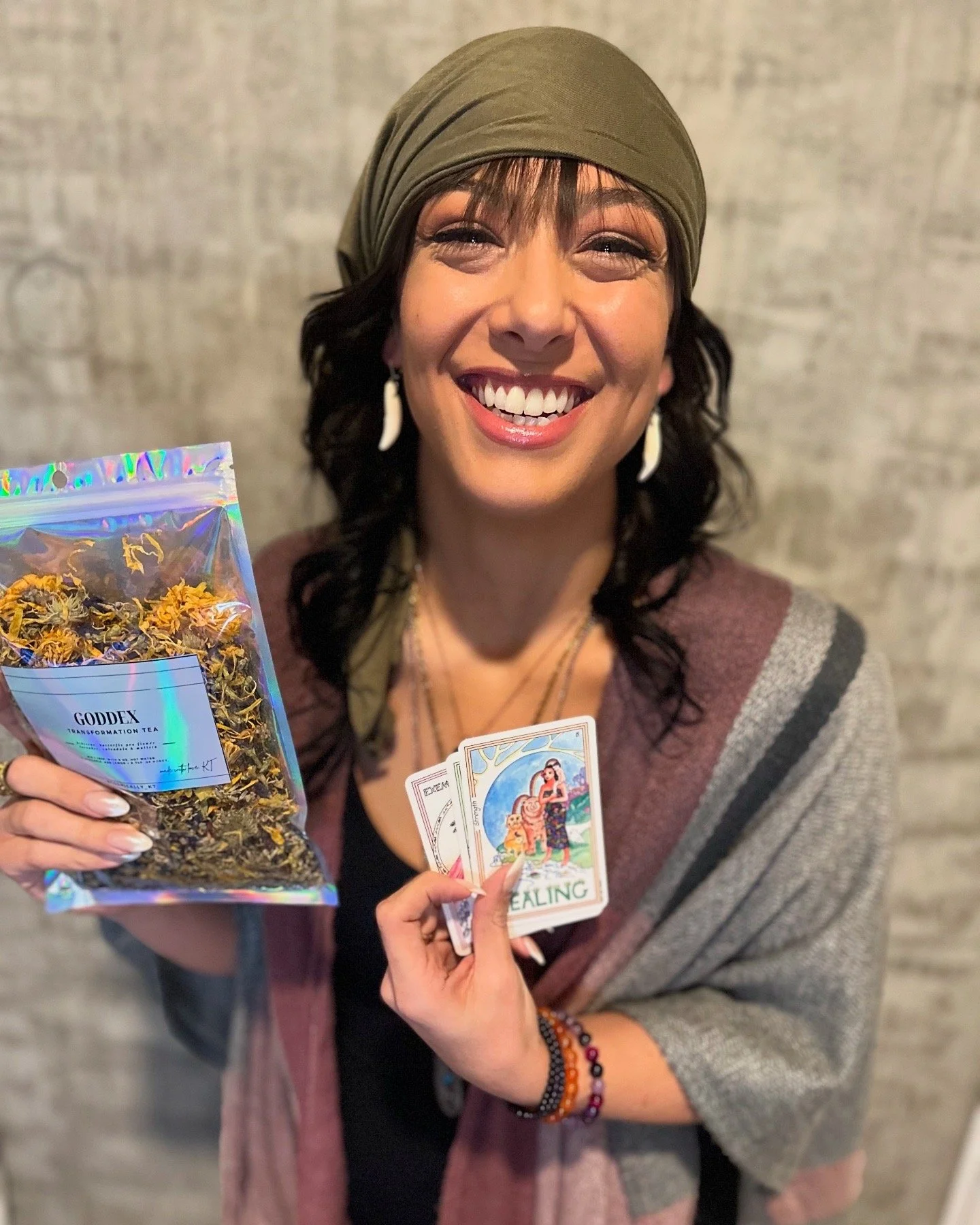 A smiling woman holding a package of herbal tea and a set of tarot or oracle cards.