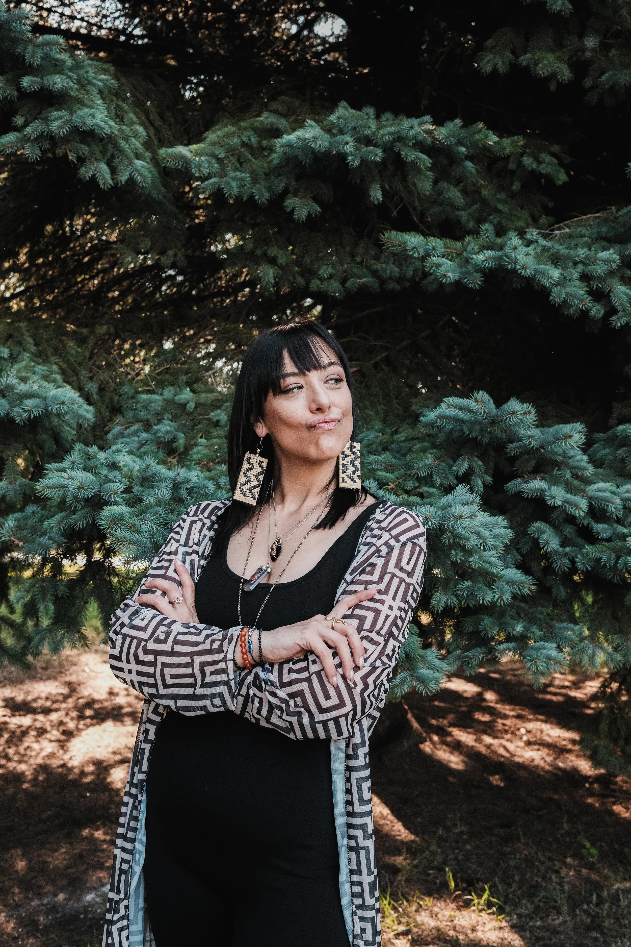 A woman with black hair and bangs posing outdoors in front of a large evergreen tree, wearing a patterned jacket over a black top, with her arms crossed and making a puckered facial expression.