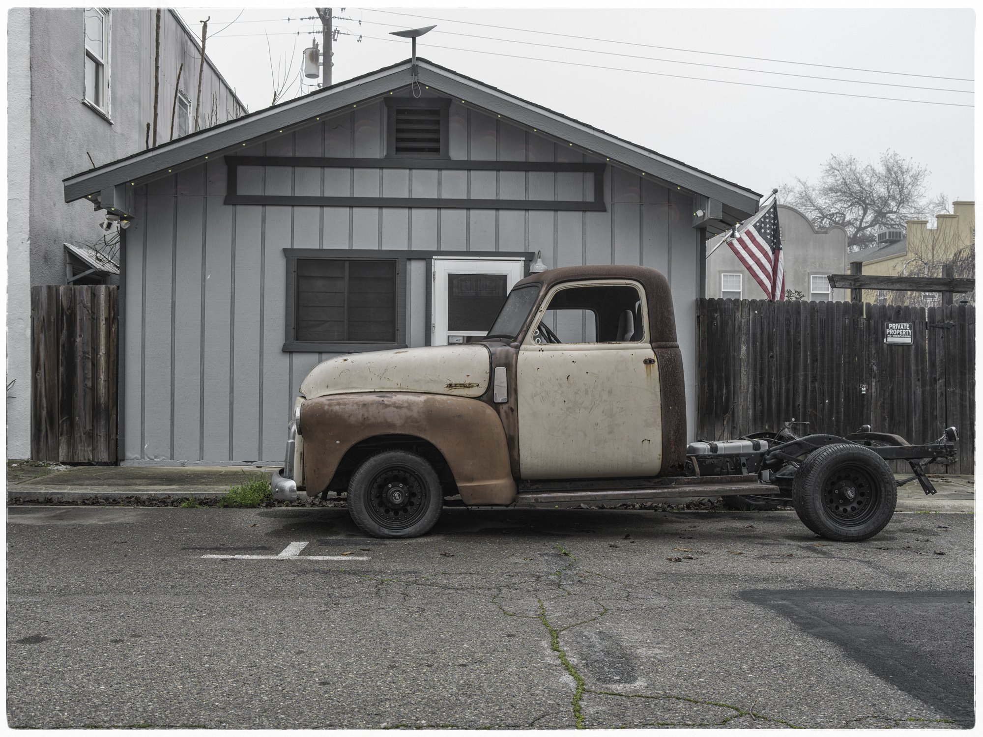 An unfinished vintage truck with rust and missing parts parked in front of a blue house with an American flag.