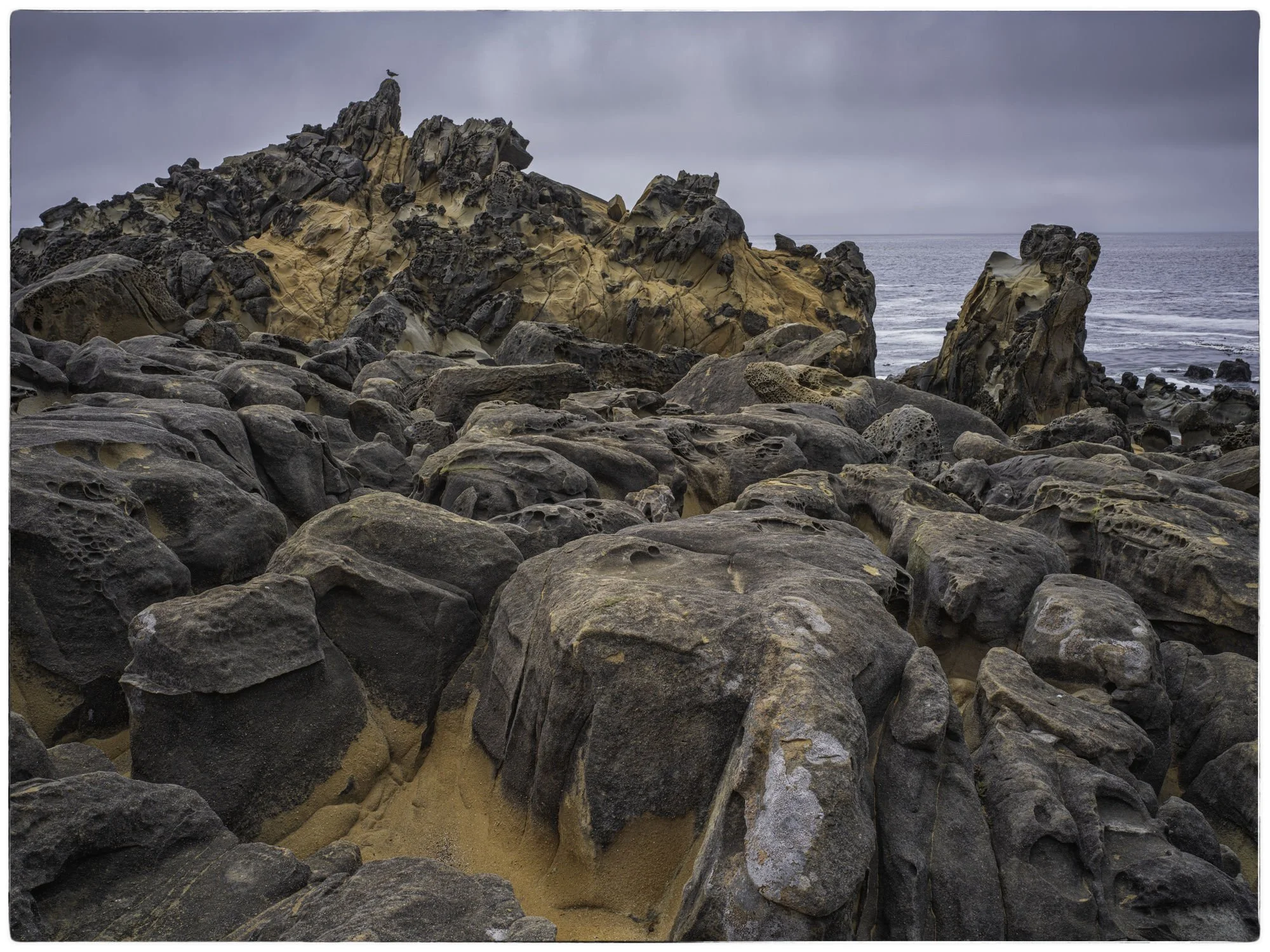 A rocky seashore with large, dark, porous rocks and jagged formations in the background, overlooking the ocean under a cloudy sky.