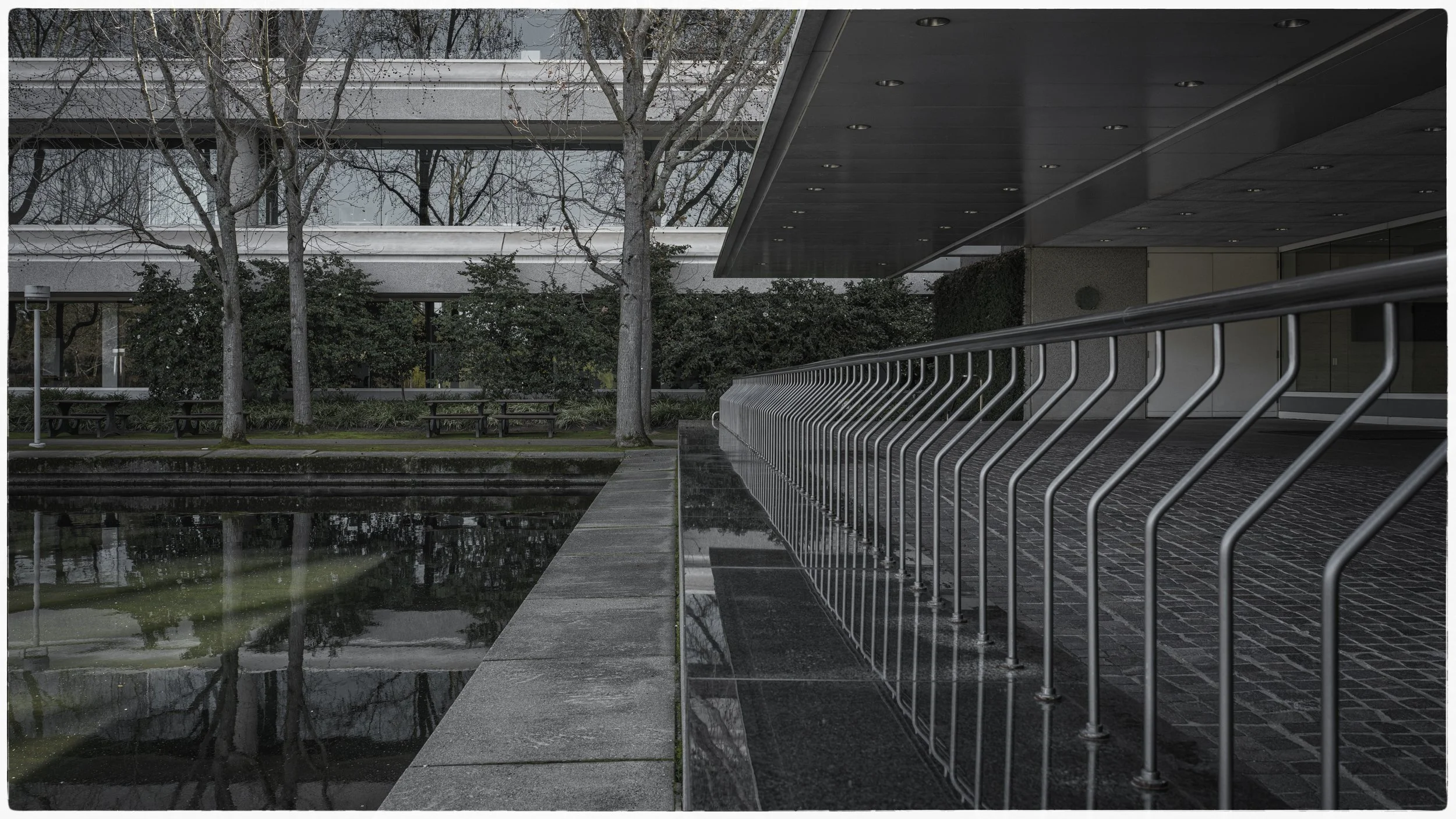 Modern building entrance with a metal railing, reflection in water, trees, and benches in the background.