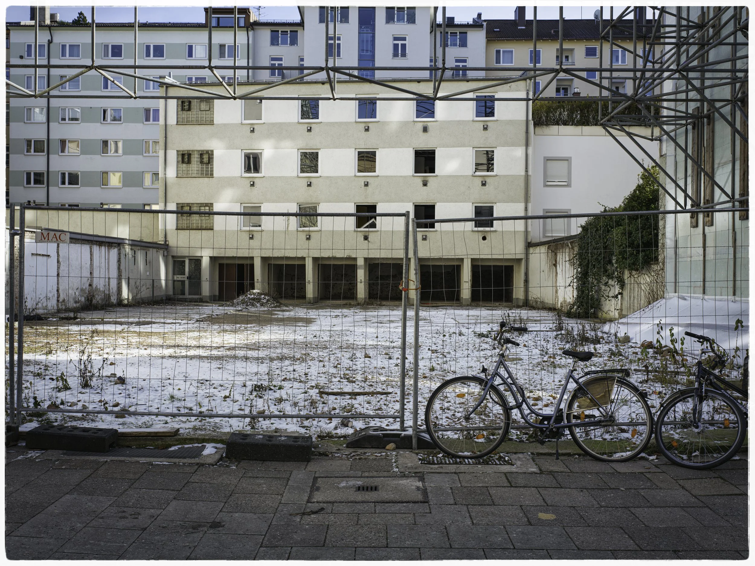 An empty urban lot enclosed by a metal fence, with a bicycle parked in front. The ground has snow and scattered leaves. Surrounding buildings are visible in the background.