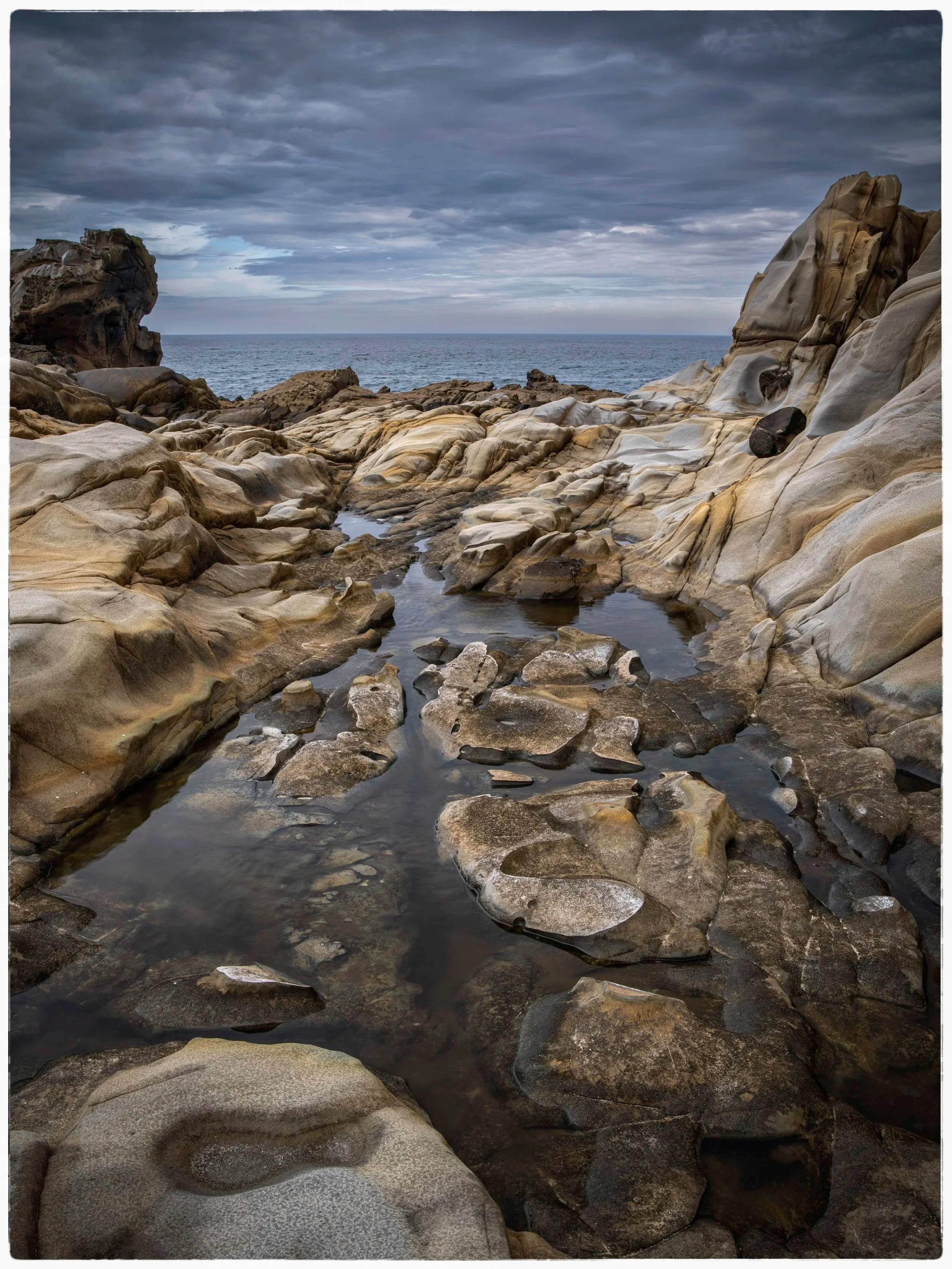 Rocky shoreline with tide pools under a cloudy sky, and the ocean in the distance.