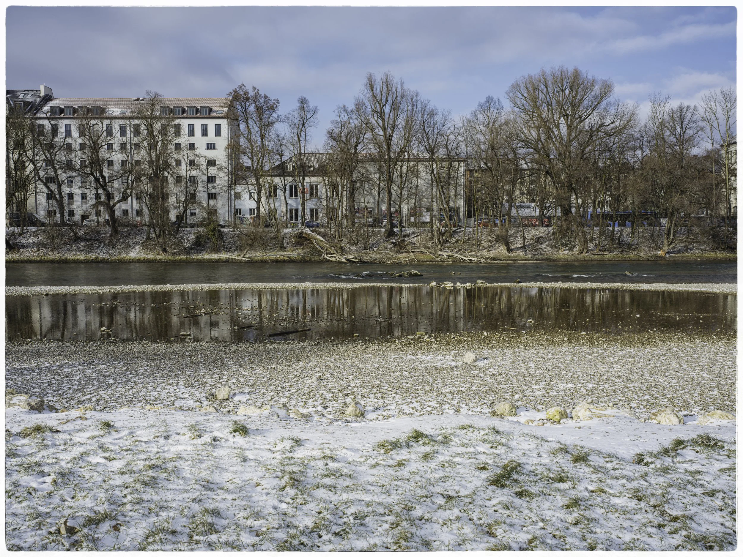 A winter scene of a river with snow-covered ground in the foreground. Bare trees line the riverbank, with apartment buildings in the background under a partly cloudy sky.