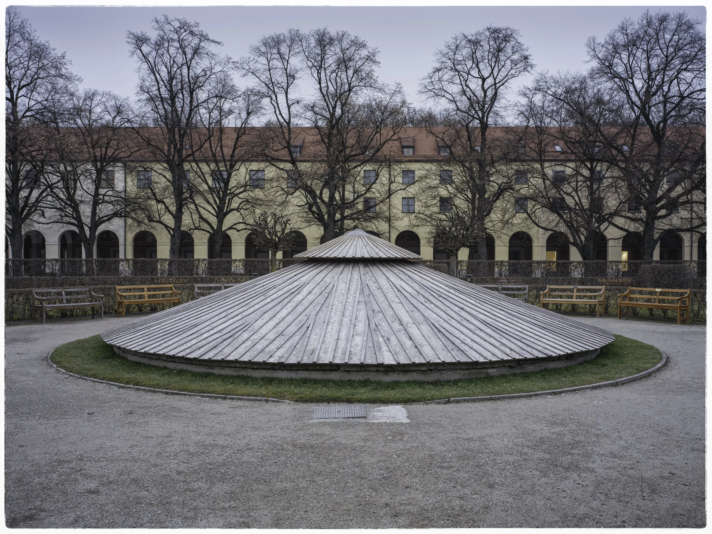 Circular wooden structure with a conical top, surrounded by benches and trees with no leaves, in a park with an apartment building in the background.