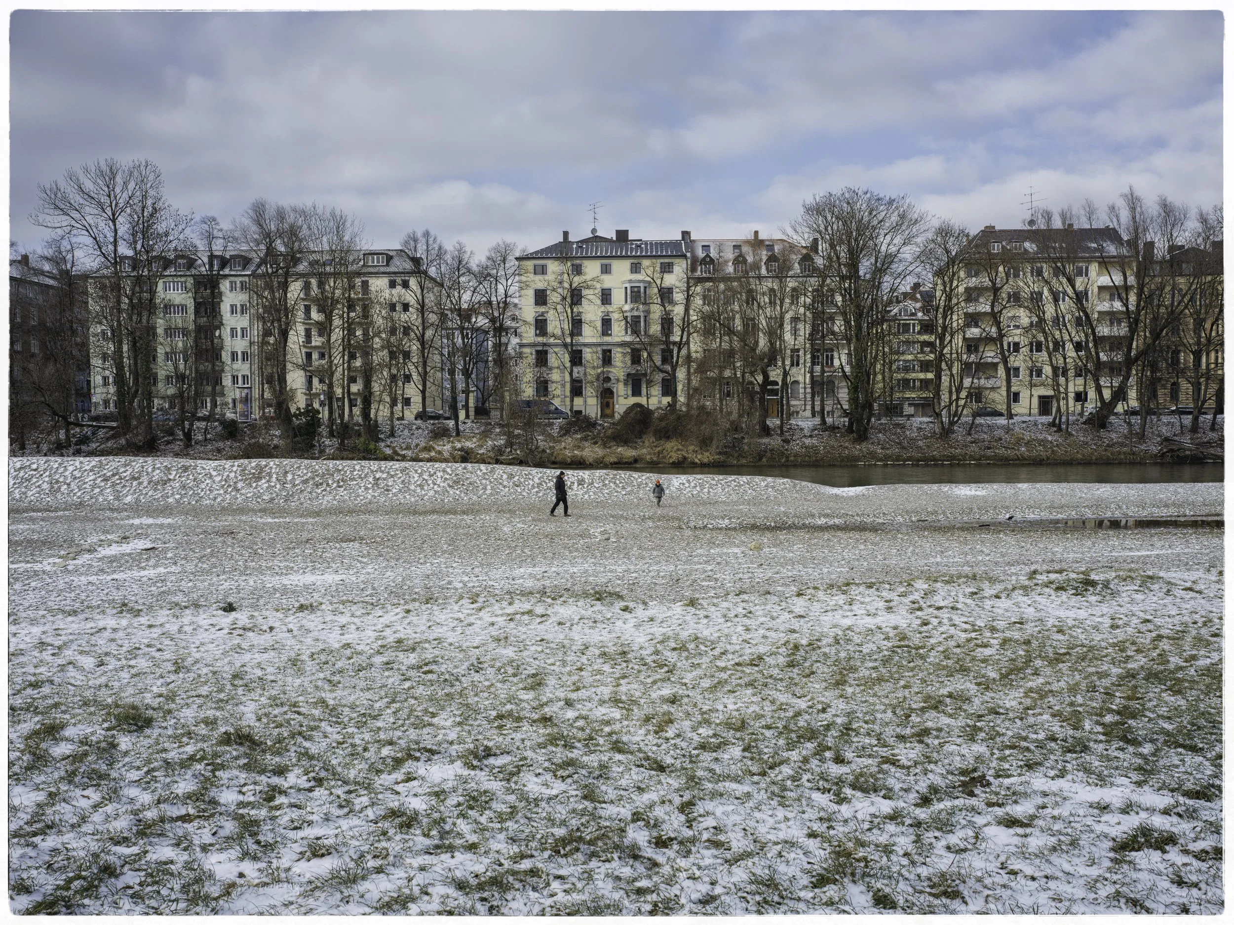A winter scene with a snow-dusted grassy field, a small river, and a row of residential buildings and leafless trees in the background under a partly cloudy sky.