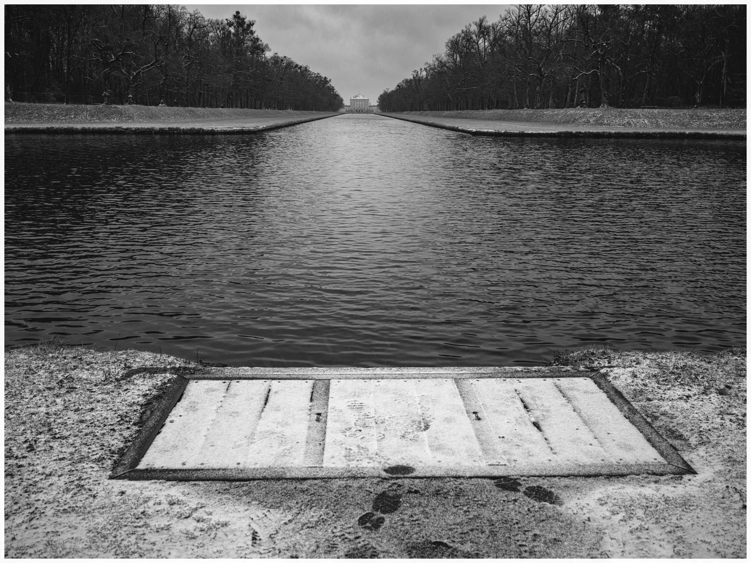 A snowy scene at a reservoir with a concrete dock in the foreground, a waterway stretching into the distance, lined with leafless trees, leading to a large building in the background.