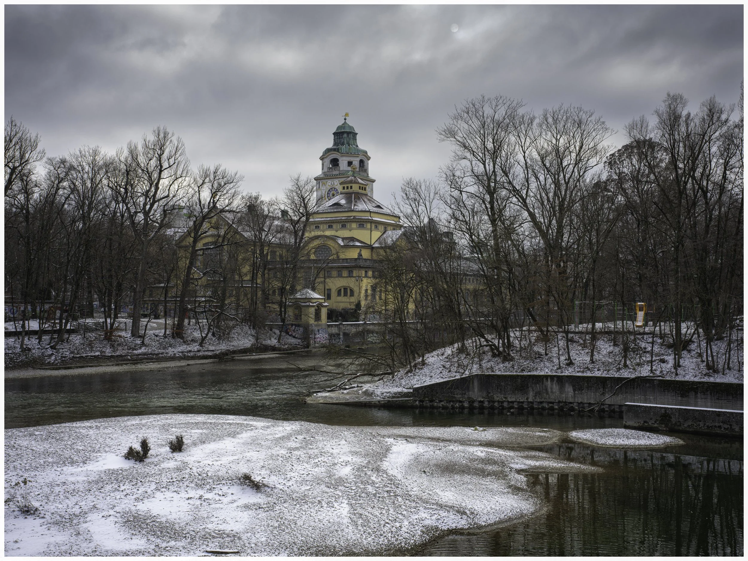 A historic yellow building with a green dome sits behind leafless trees by a river on a cloudy winter day. Snow covers the ground and parts of the river.