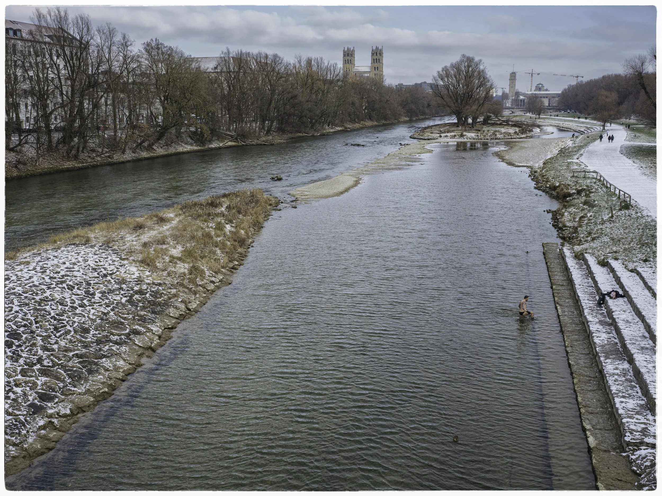 View of a river with some people swimming and walking along the snowy banks on a partly cloudy day, with buildings and trees in the background.