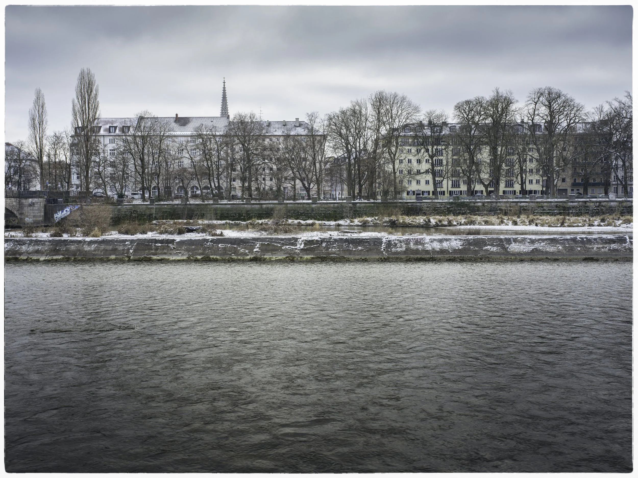 A river with a cityscape on the opposite bank, snow-covered ground and leafless trees, under cloudy sky.