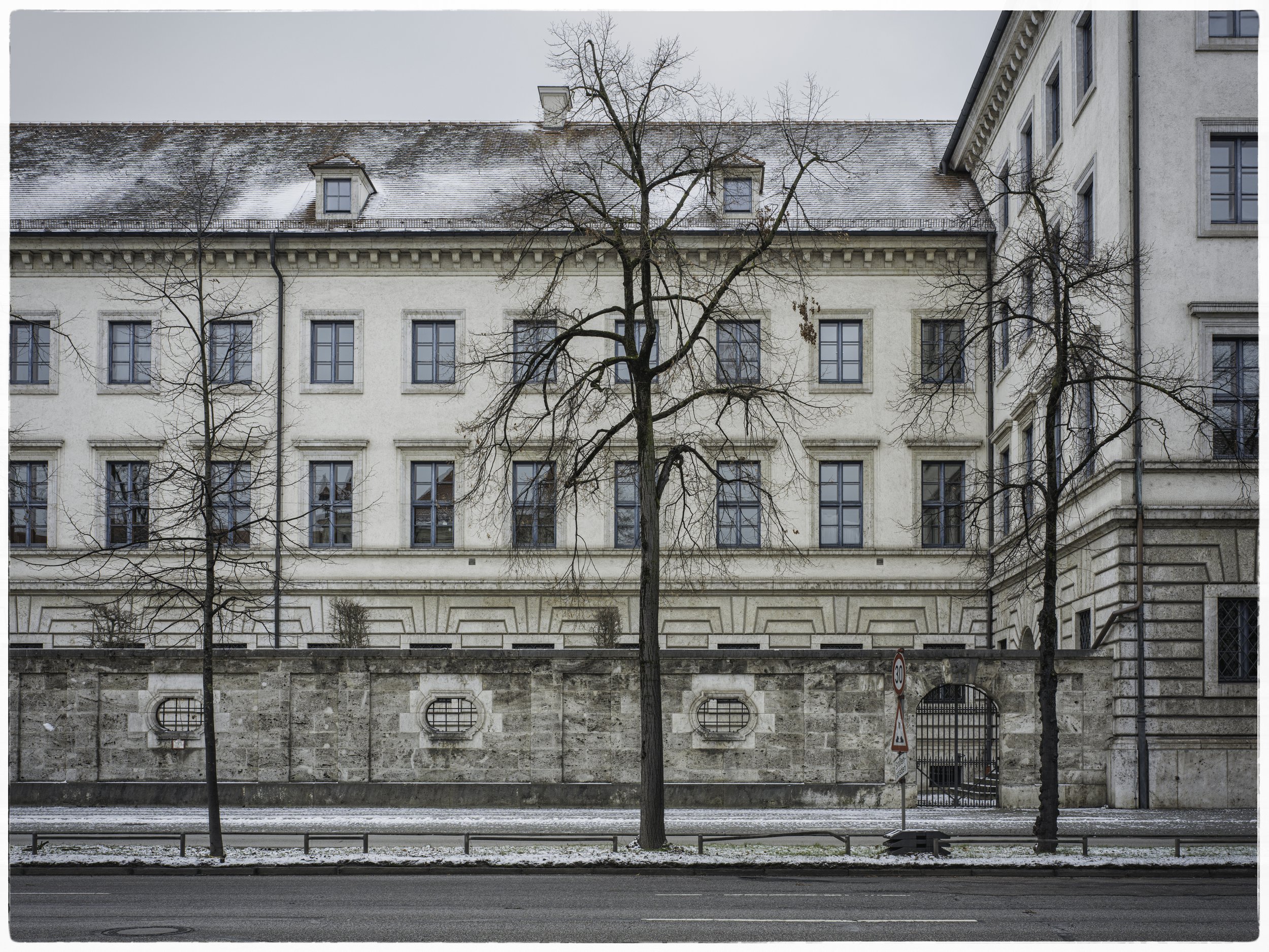 A street view of a large, historic building with multiple windows, a stone wall, and leafless trees in front, on a cold winter day.