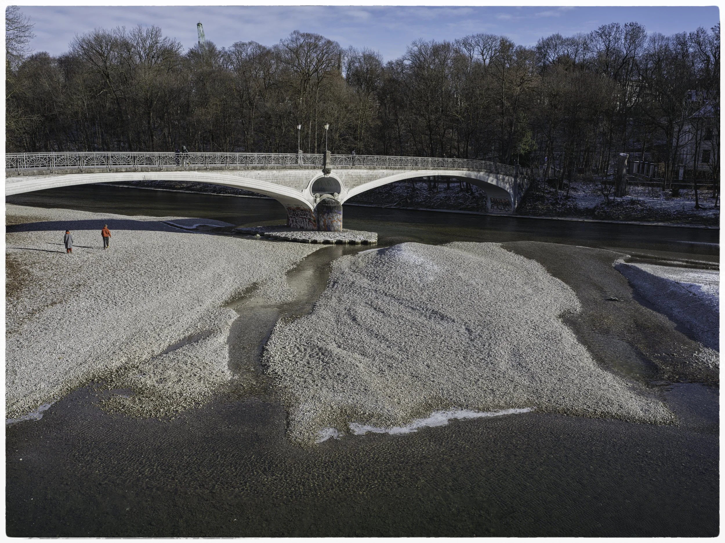 A white bridge crosses over a river in a cold, winter landscape. There are two people walking on the snow-covered riverbank near the water.