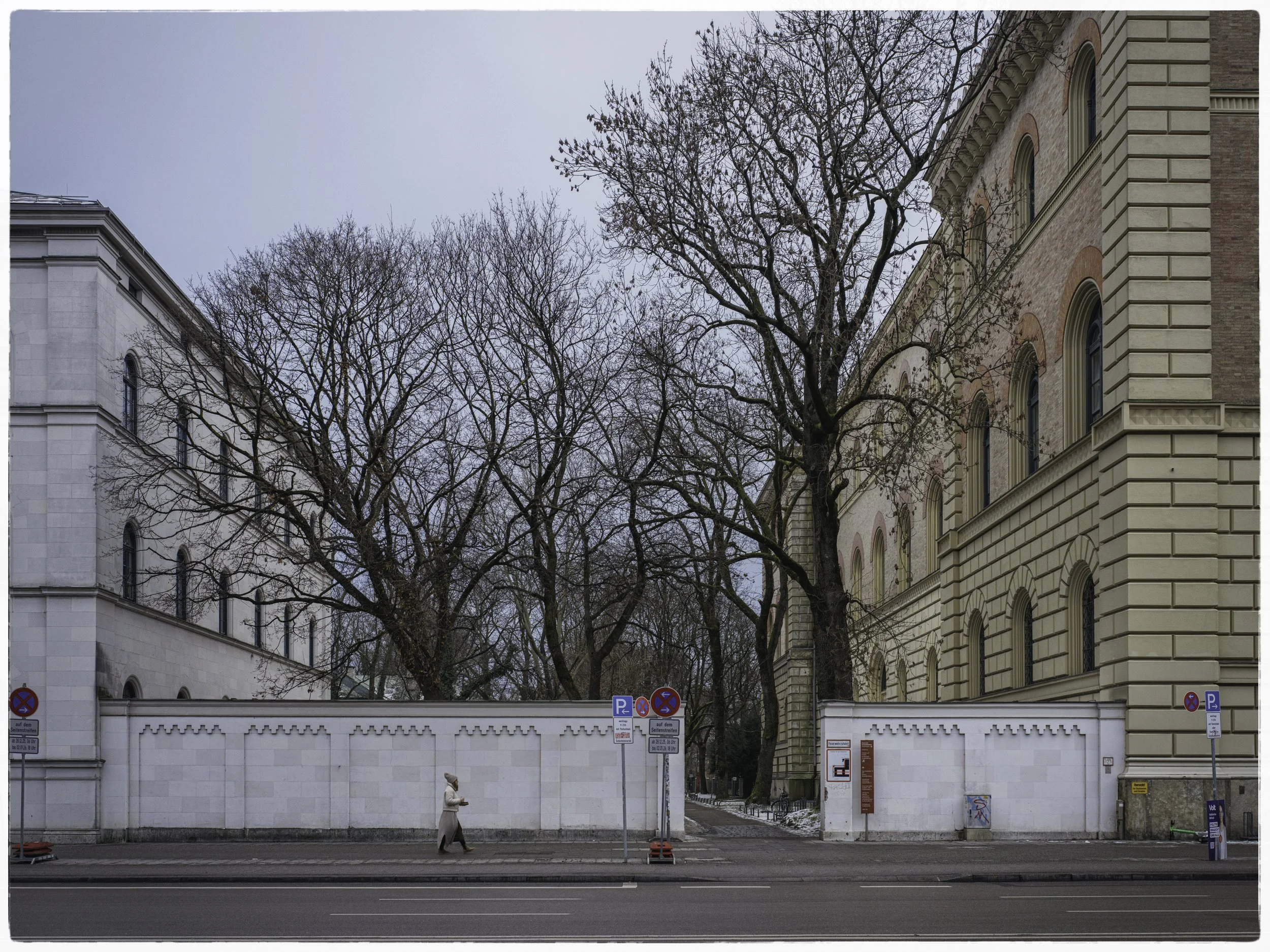A street scene with a woman walking along the sidewalk, a white wall with decorative top, and large trees with no leaves. Behind the wall are old buildings with classical architecture, windows, and brick facades. Several parking signs are visible alo