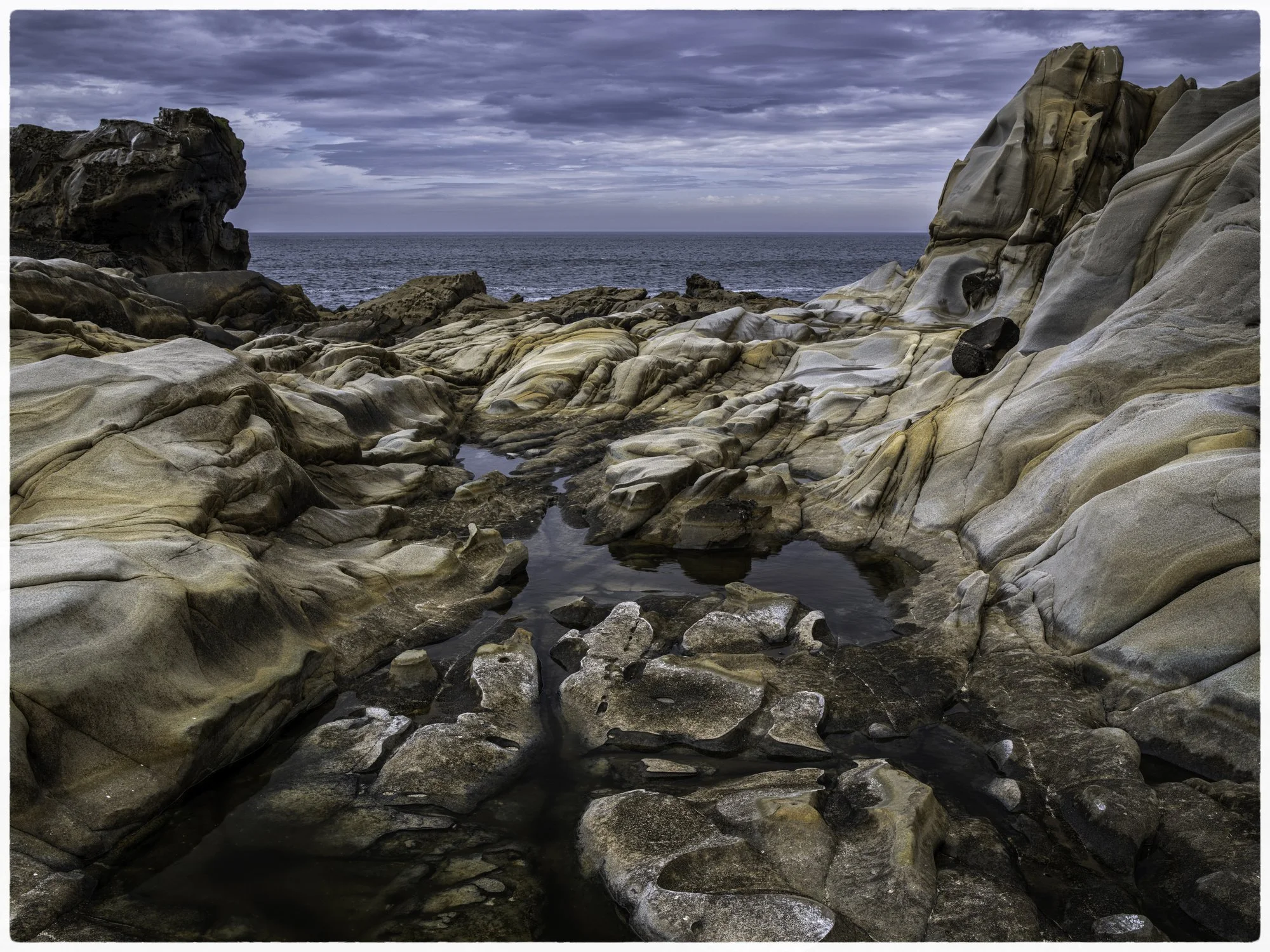 Rocky shoreline with tide pools and ocean in the background under cloudy sky.
