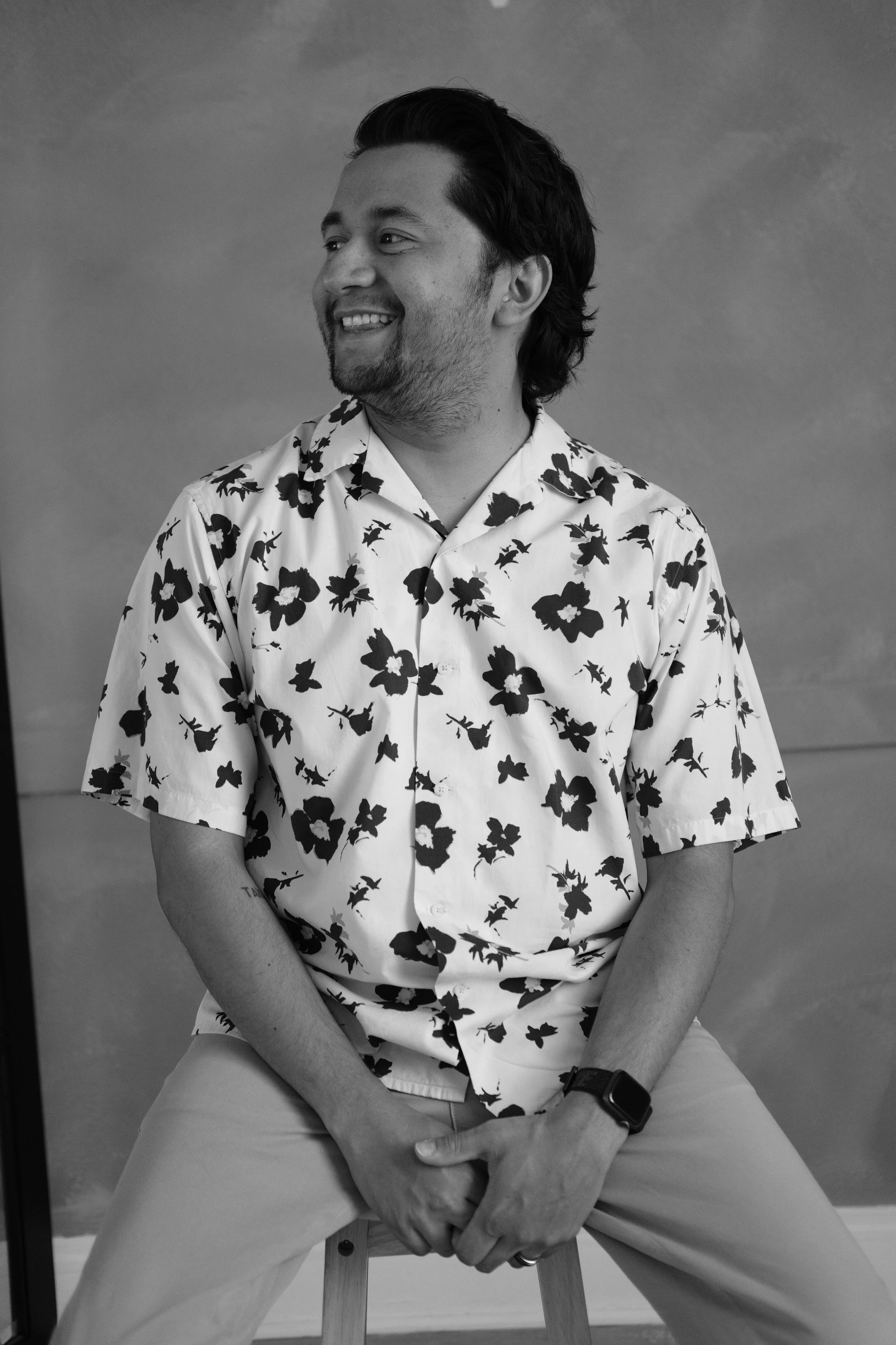 A smiling man with dark hair, beard, and mustache, sitting on a stool, wearing a floral patterned shirt and light-colored pants, in front of a plain backdrop.