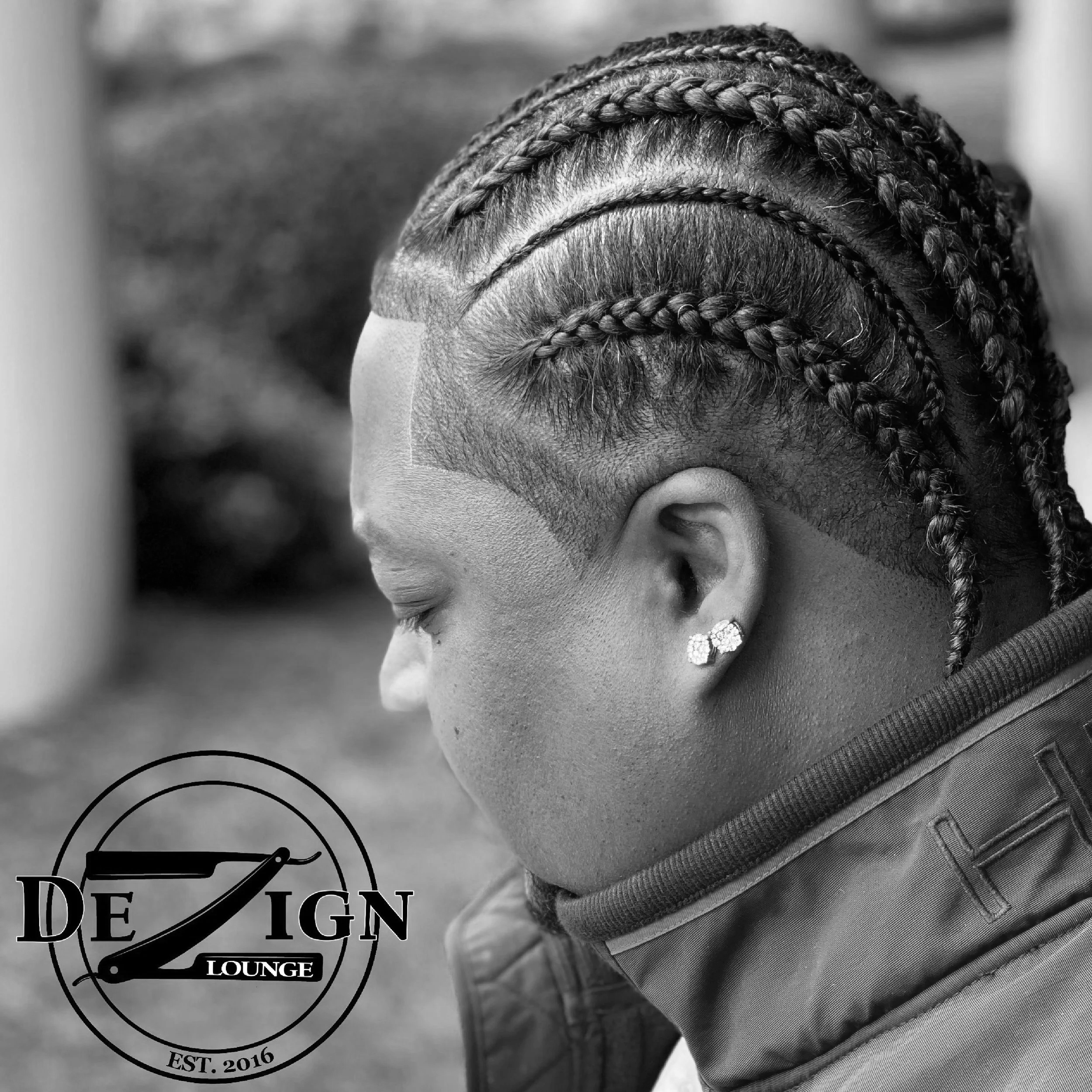 Side view of a woman with intricate braided hairstyle, shaved sides, and wearing pearl earrings. She is outdoors with a blurred background.