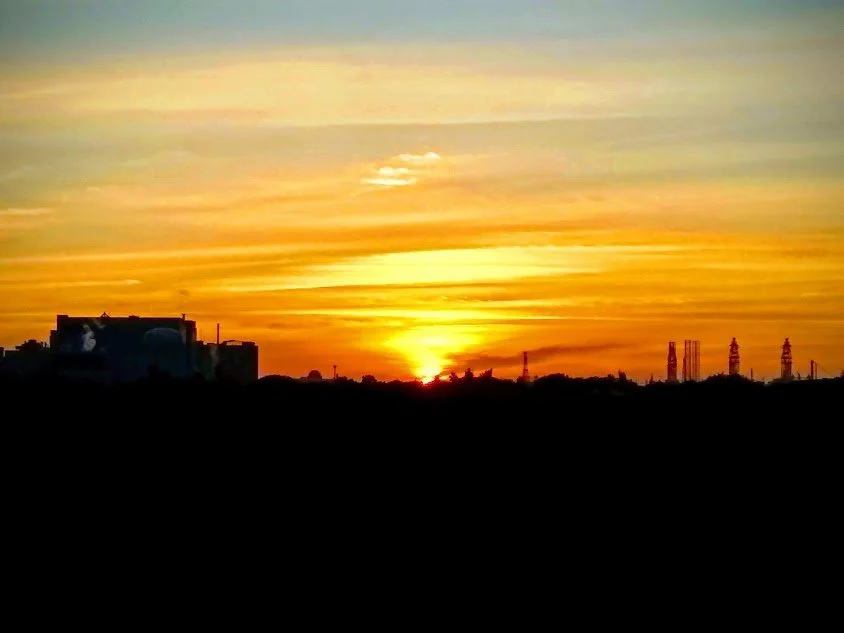 Sunset over a horizon with a silhouette of buildings and communication towers, with colorful sky in shades of yellow, orange, and blue.