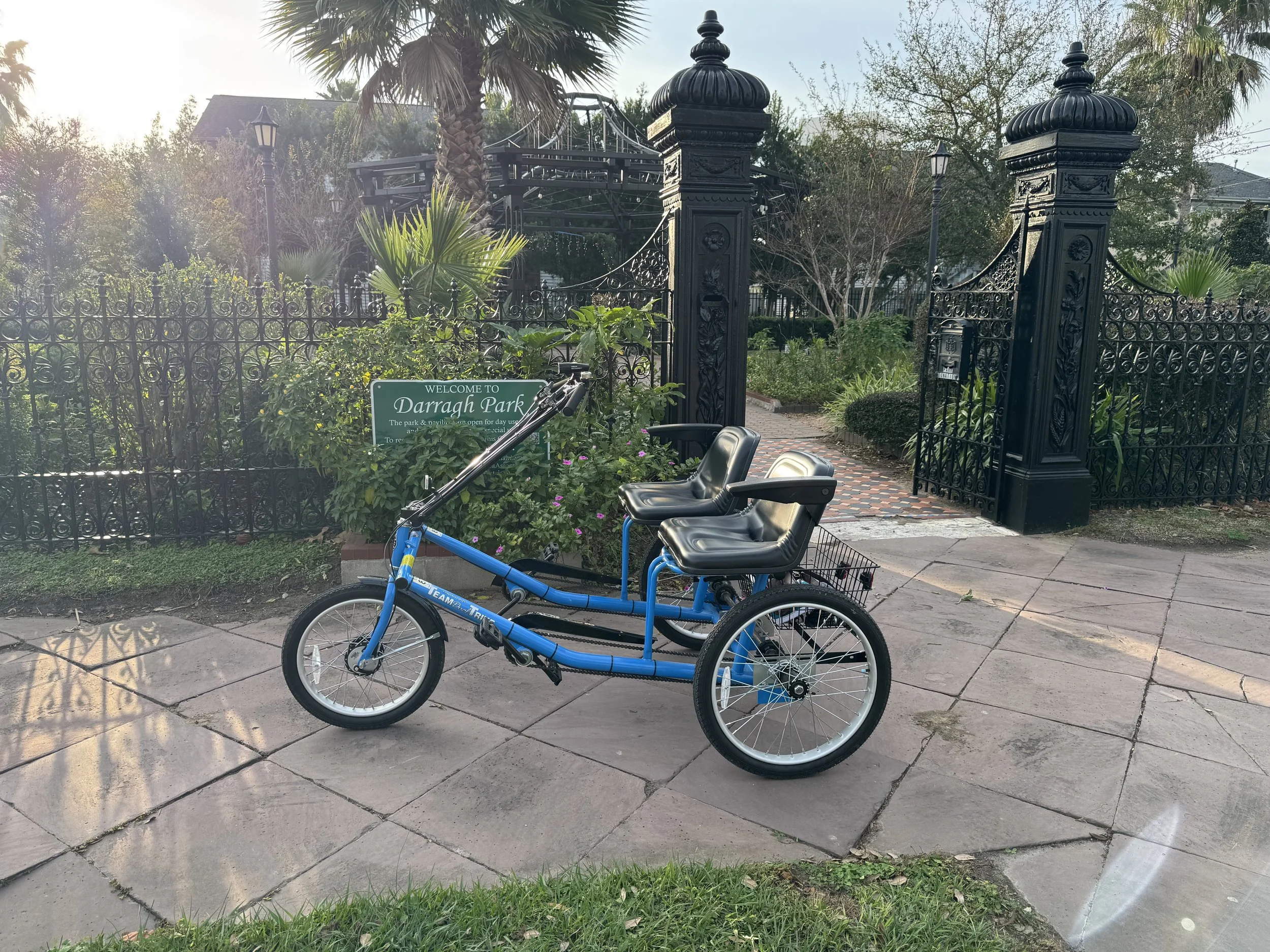 A blue four-seater bike with black seats parked on a sidewalk in front of a black wrought-iron gate and fence at Darragh Park. There are green plants and a sign that reads "Welcome to Darragh Park." Palm trees and houses are visible in the background.