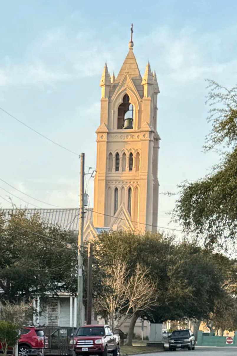 A tall church steeple with pointed spires and arched windows, seen above trees and parked cars on a suburban street during sunset.