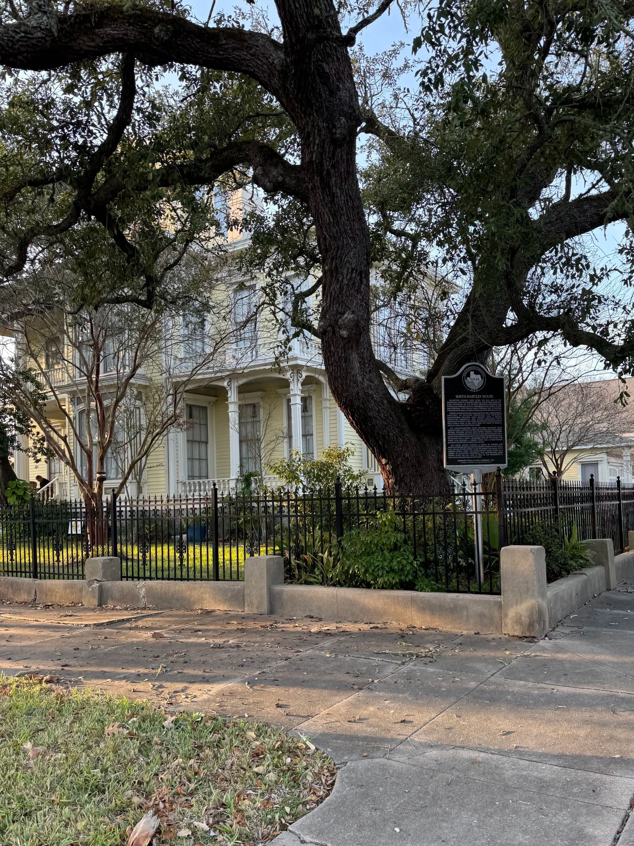 A historic yellow house with white trim, surrounded by a black fence, with a large tree in the front yard and a historical marker sign near the tree.