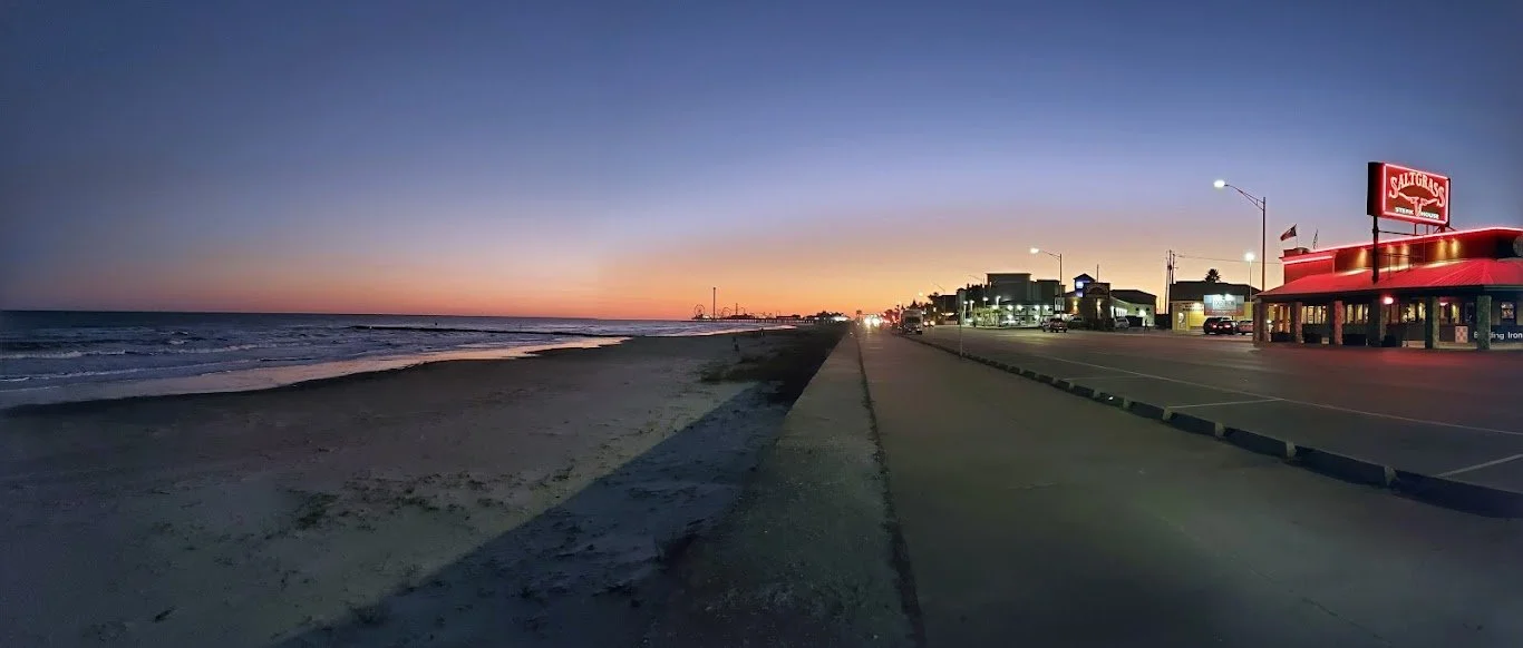 Sunset view of the beach and ocean on the left, with a boardwalk and a row of buildings, including the Saltgrass Steak House, on the right in a small coastal town.