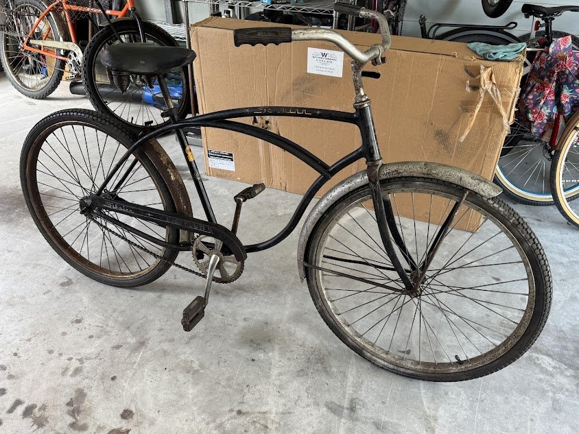 An old black bicycle with rust on the wheels and frame, parked on a concrete floor with other bicycles and a large cardboard box in the background.