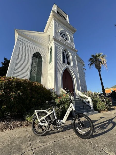 A white church with a tall steeple, arched windows, and a red door, with a bicycle parked in front and a palm tree nearby on a clear blue day.
