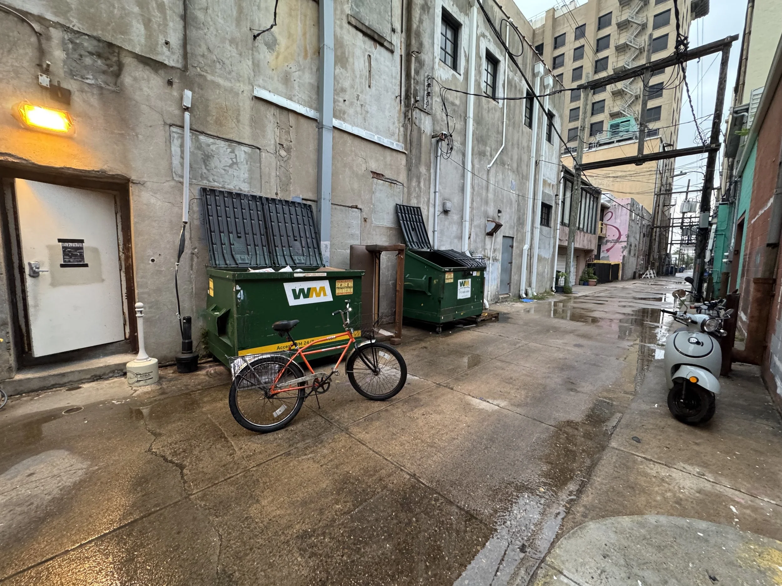 Back alley with wet concrete ground, two green dumpsters, a parked white scooter, and a red bicycle leaning against one dumpster. The alley is surrounded by aged buildings with exposed utilities and wires, and a distant taller building is visible.