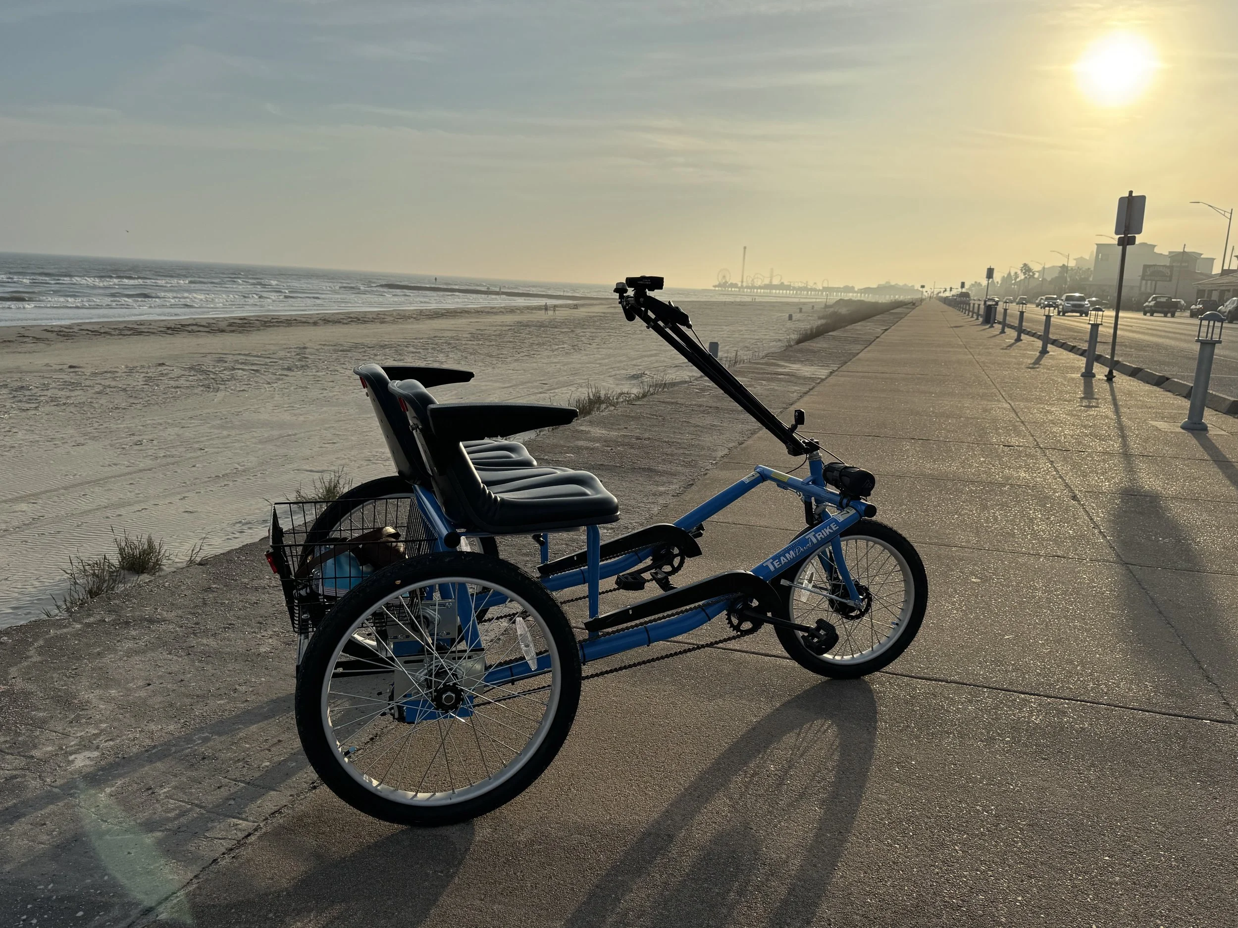 Blue pedal-powered bike with three seats and a basket on the back, parked on a beachside promenade at sunset.