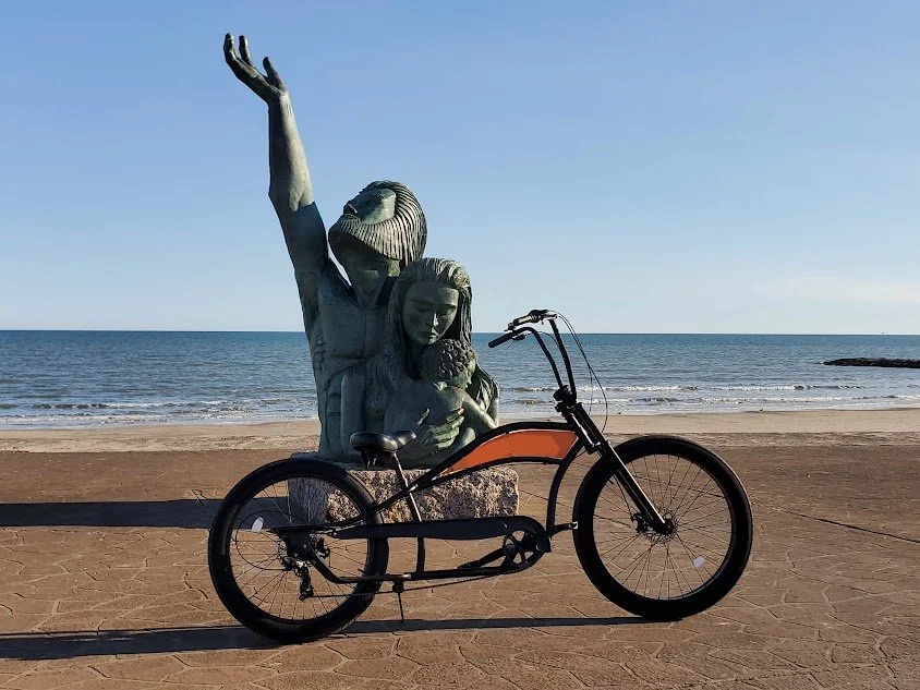 Statue of a woman with one arm raised, holding a young girl, with the ocean in the background, and a bicycle in front of the statue on the beach.