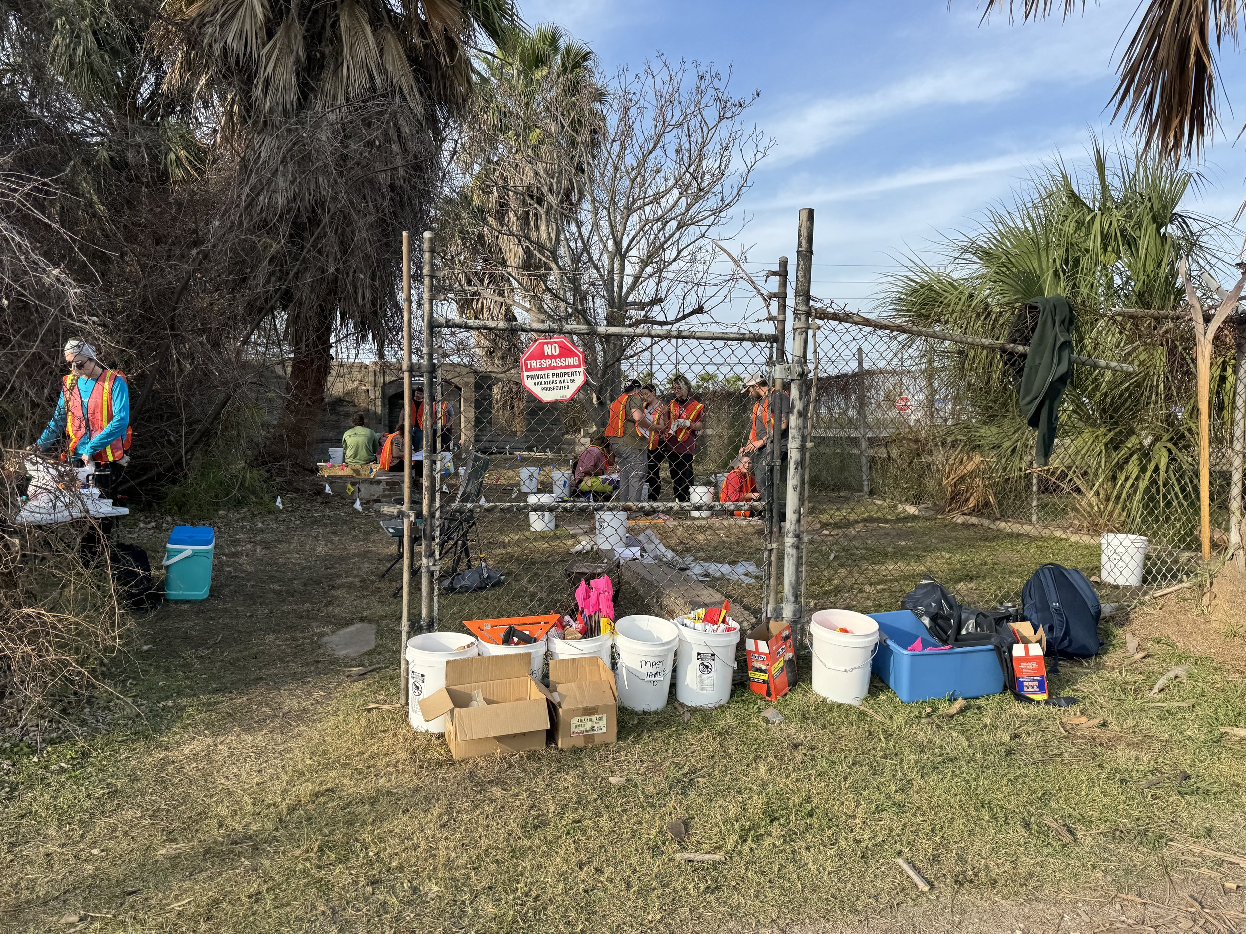 Several people wearing safety vests working inside a fenced area with tools, buckets, and boxes outside the gate. Some trees and bushes are visible around the area.