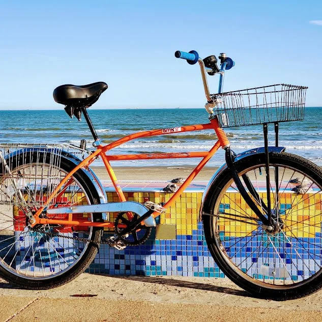 Orange bicycle with a front basket parked at the beach, with ocean and blue sky in the background.
