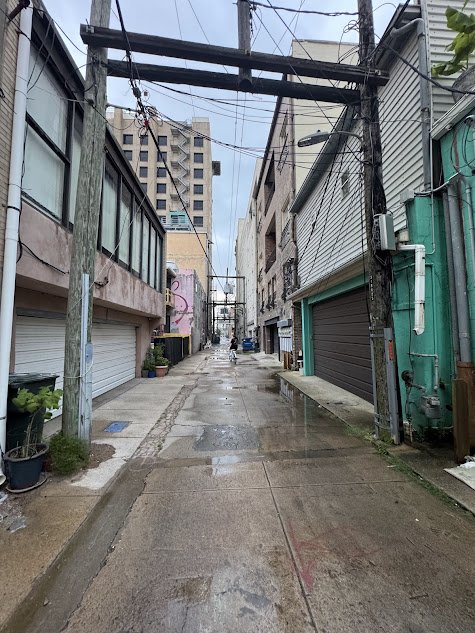 Narrow alleyway in an urban area with buildings on both sides, overhead electrical wires, and a wet sidewalk.