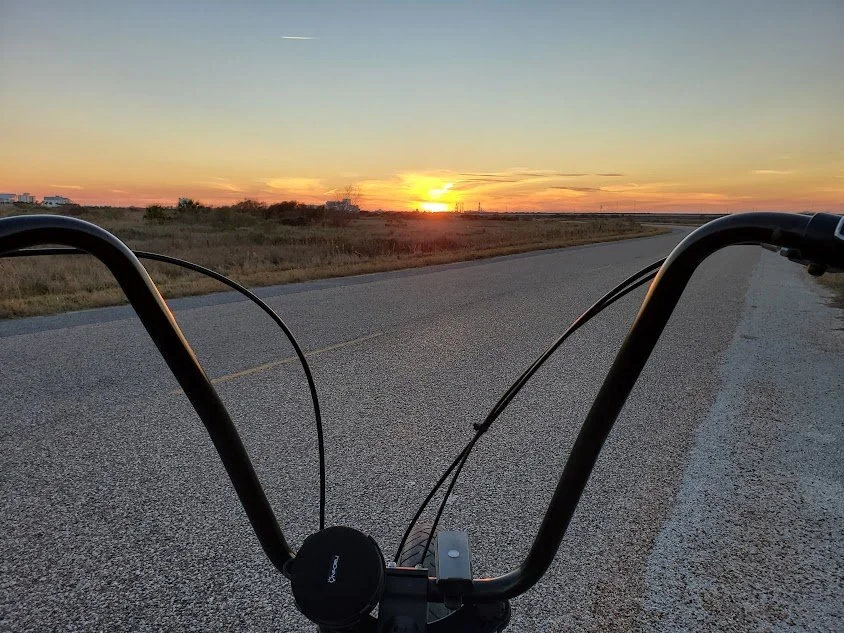 View of an open road at sunset from the perspective of someone on a bicycle, with handlebars and part of the bike visible in the foreground.