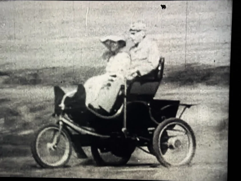 An old black-and-white photo of a small, vintage race car with a driver wearing a helmet, moving on a dirt track in front of a plain wall.